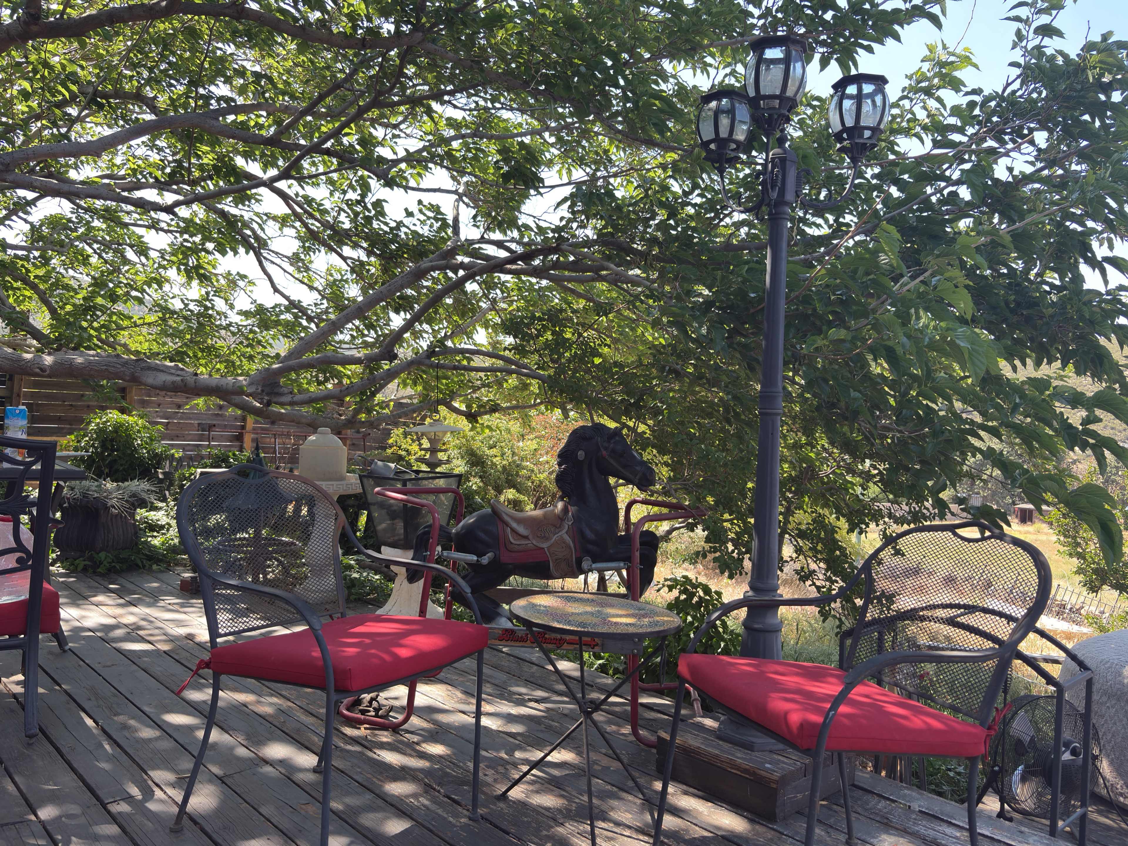 A wooden deck features three chairs with red cushions, a small table, and a horse statue under a tree.