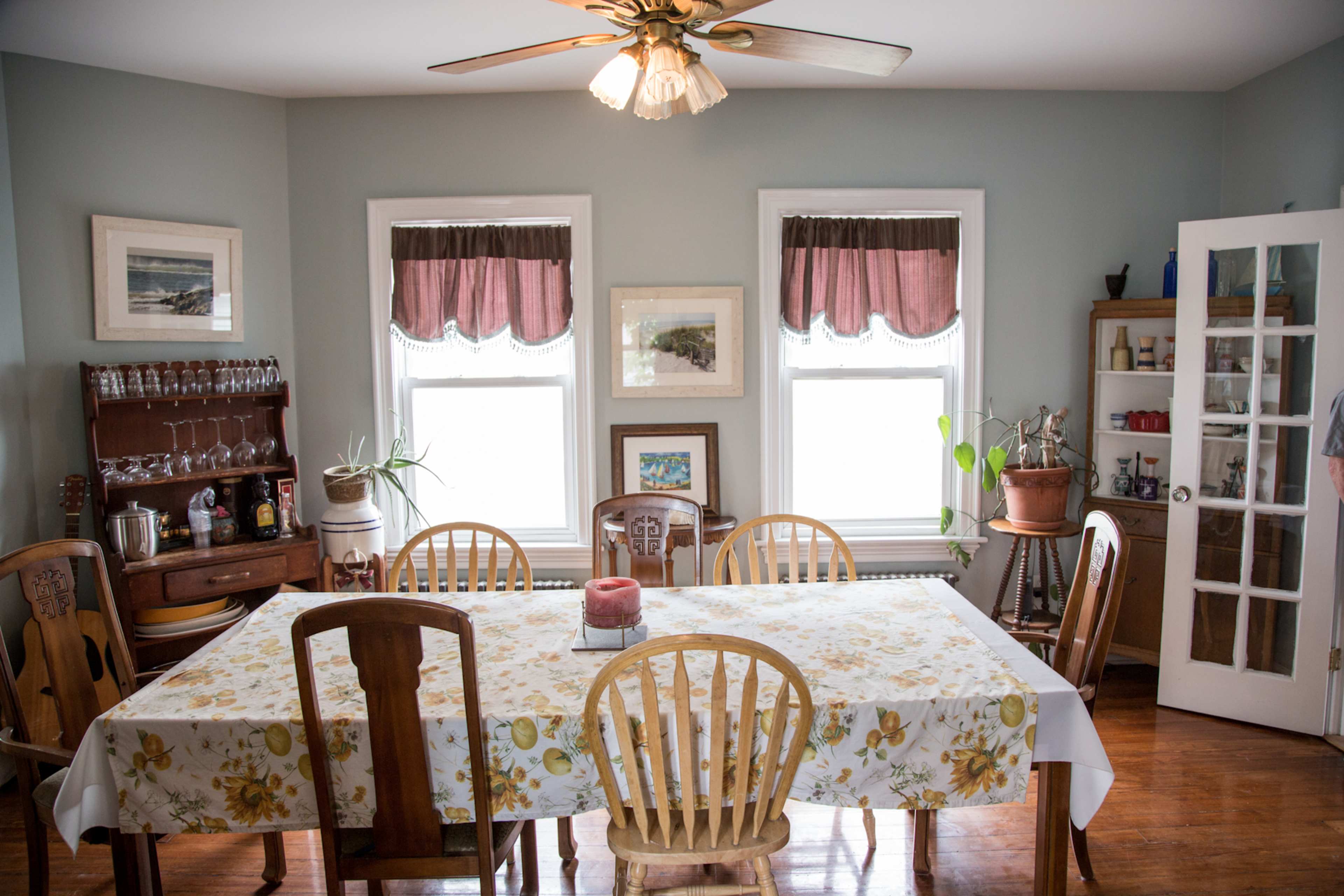 A dining room features a large table surrounded by wooden chairs, with windows adorned by roman shades and a sideboard displaying glassware.