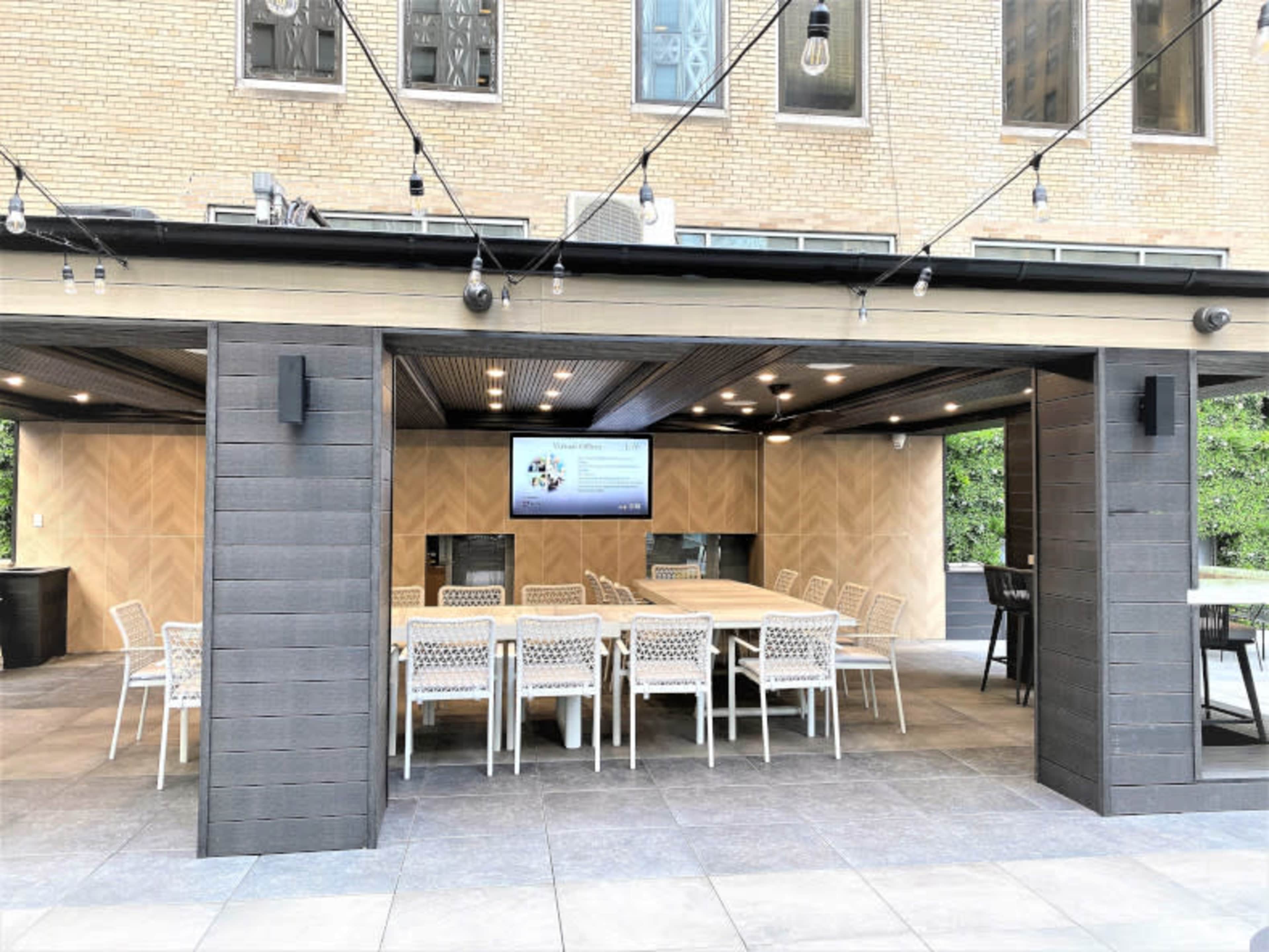 An outdoor dining area with a large table surrounded by chairs, and a screen mounted on the wall, is set beneath string lights.