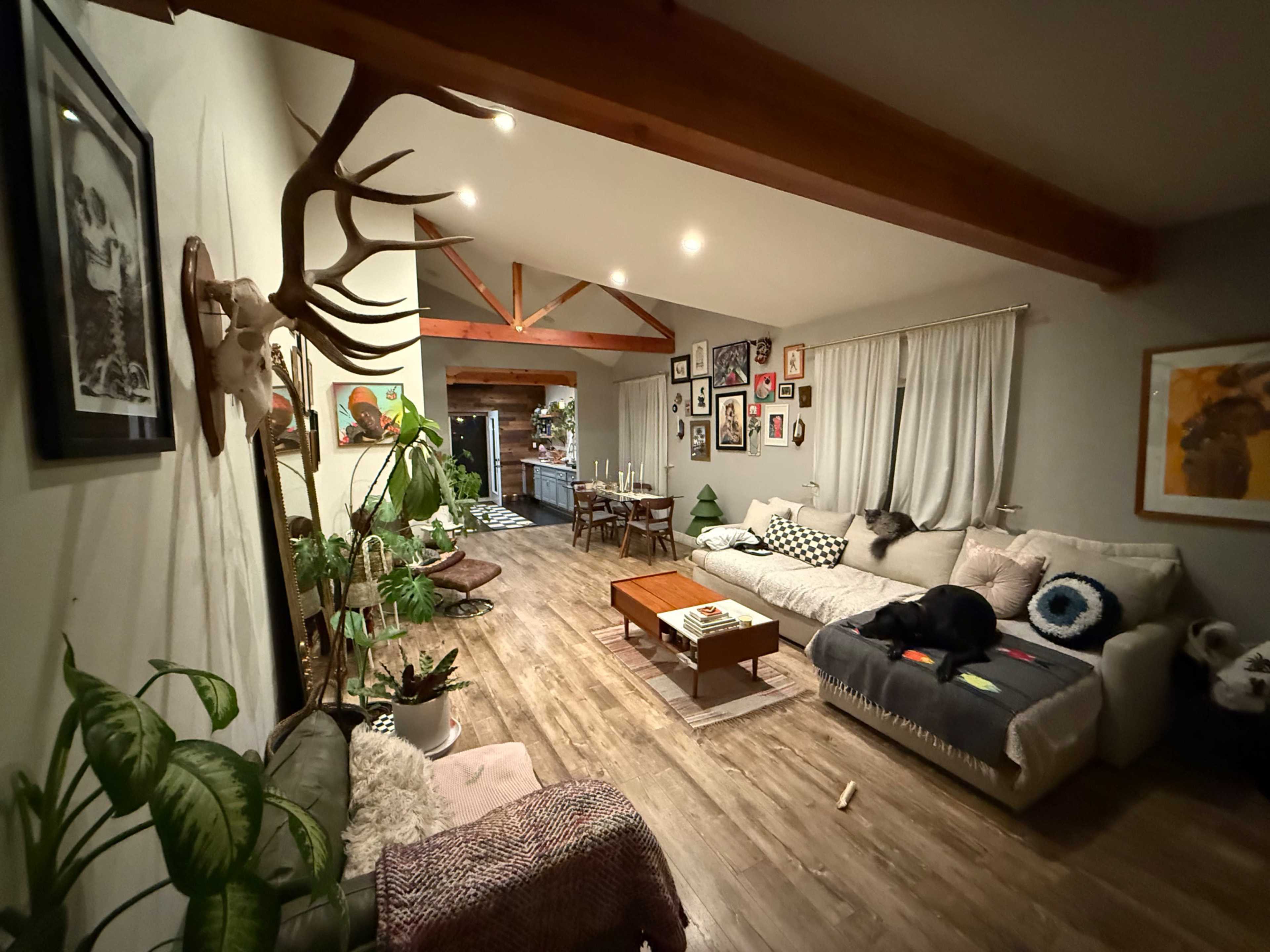 The image shows a cozy living room featuring a sectional couch, a coffee table, various wall art, and a dining area visible in the background.