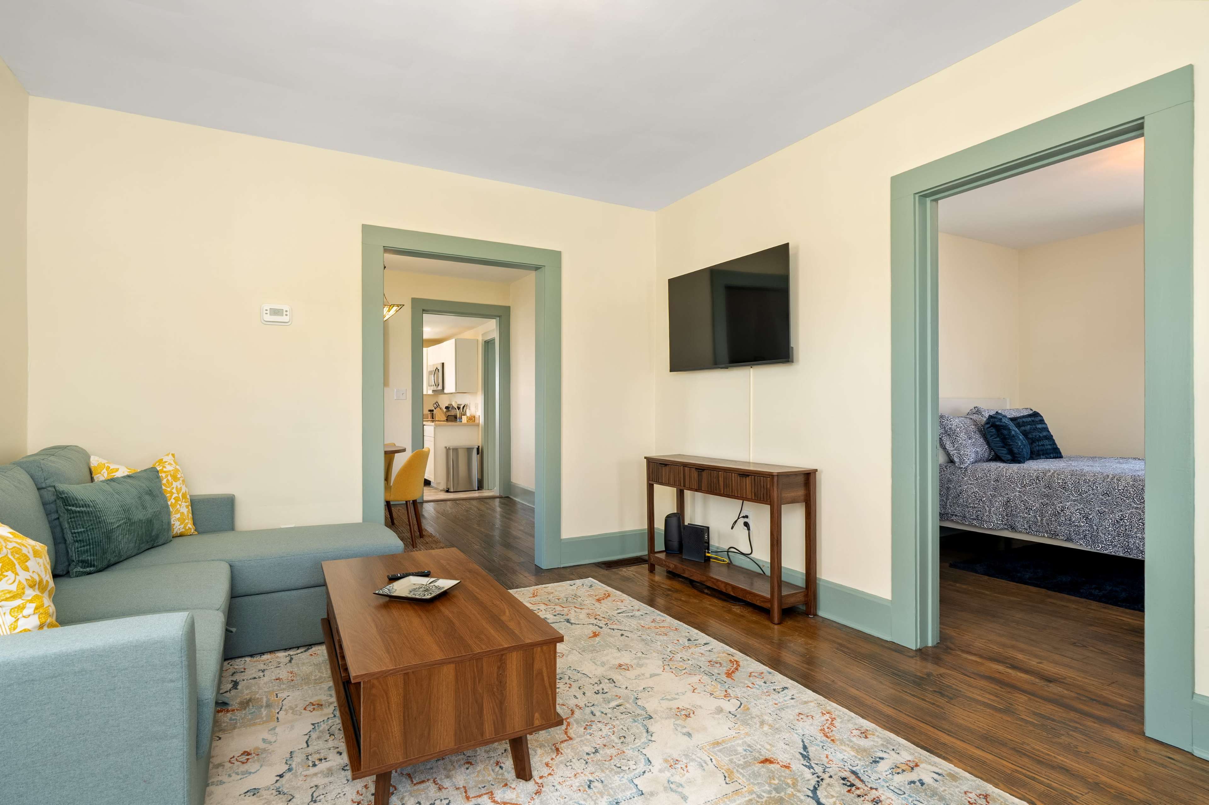 The image shows a cozy living space featuring a green sofa, a wooden coffee table, and a television, with doorways leading to another room and a bedroom in the background.
