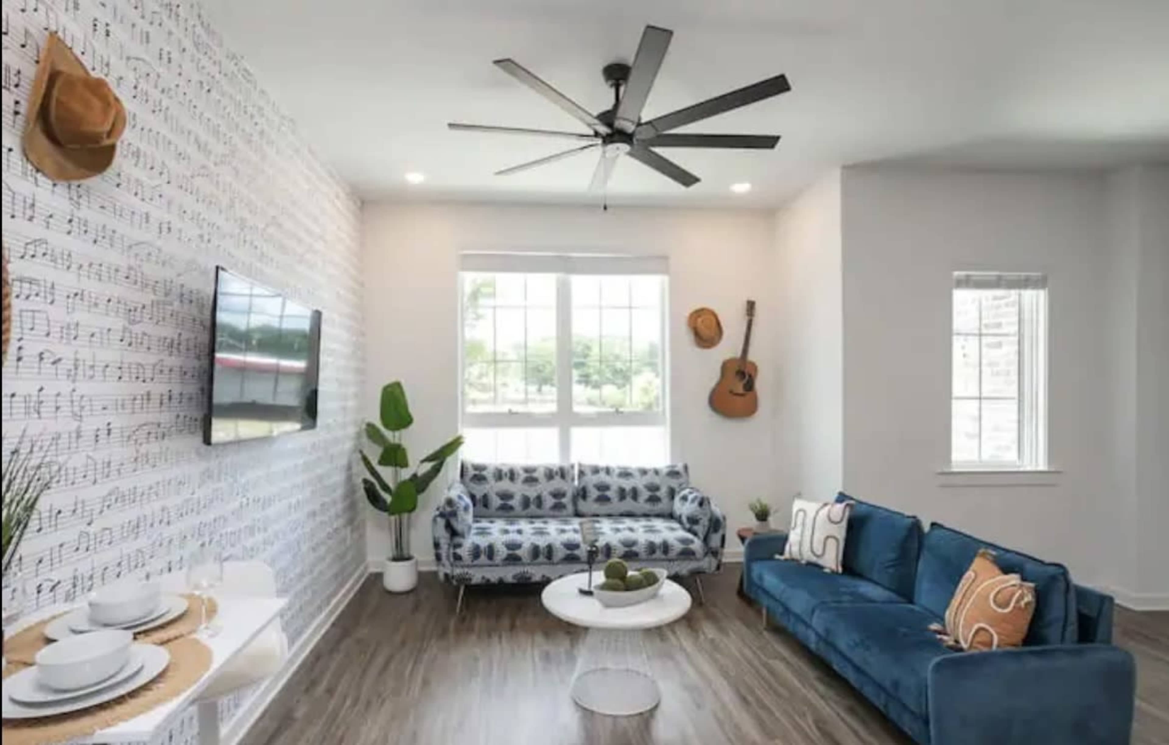 A modern living room features a blue couch, a patterned sofa, a flat-screen TV, and a guitar hanging on the wall, with a large window allowing natural light.