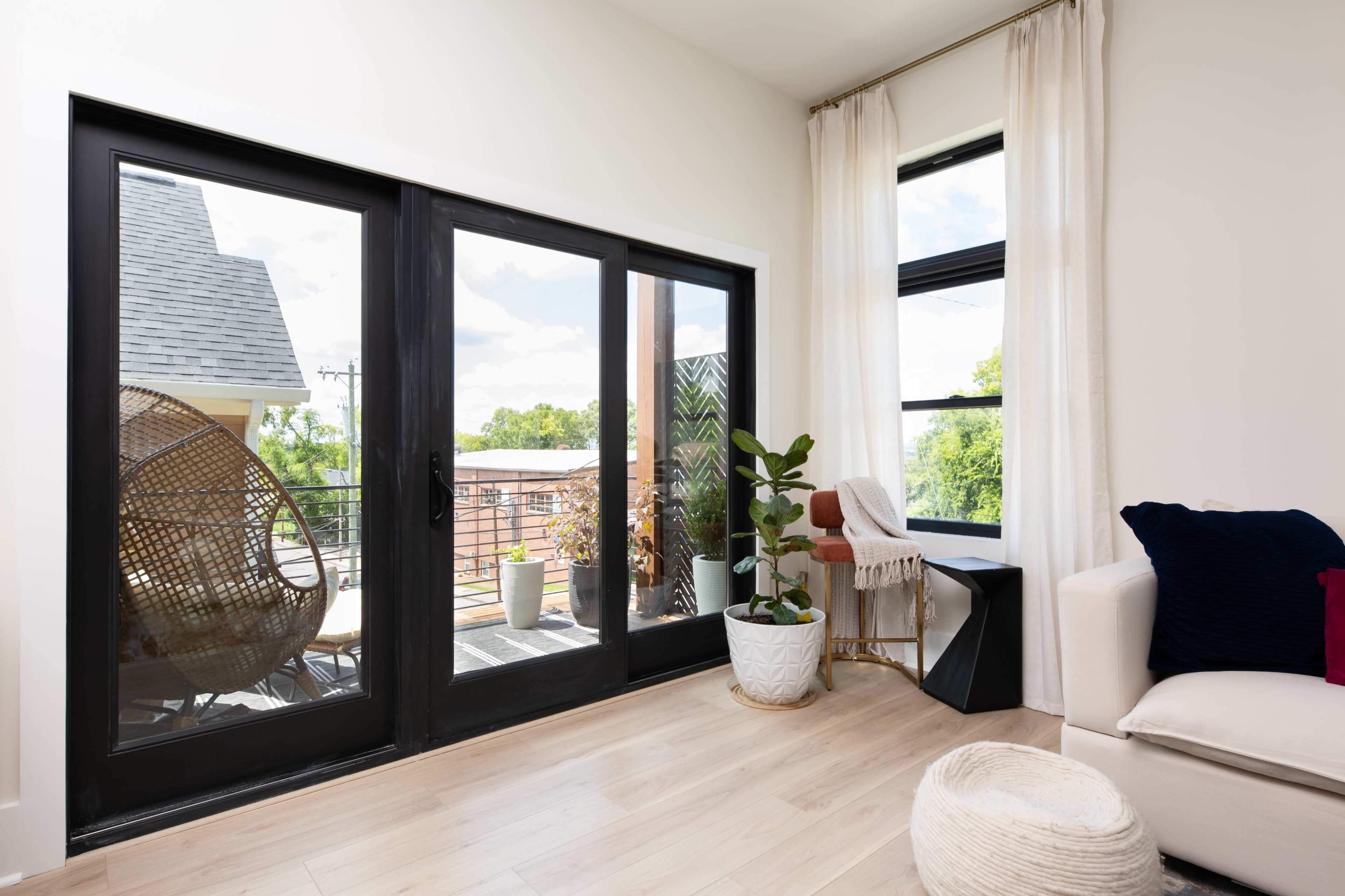 The image shows a bright interior room with large black-framed windows, a plant in a white pot, and a cozy seating area featuring a light-colored couch and a decorative throw pillow.