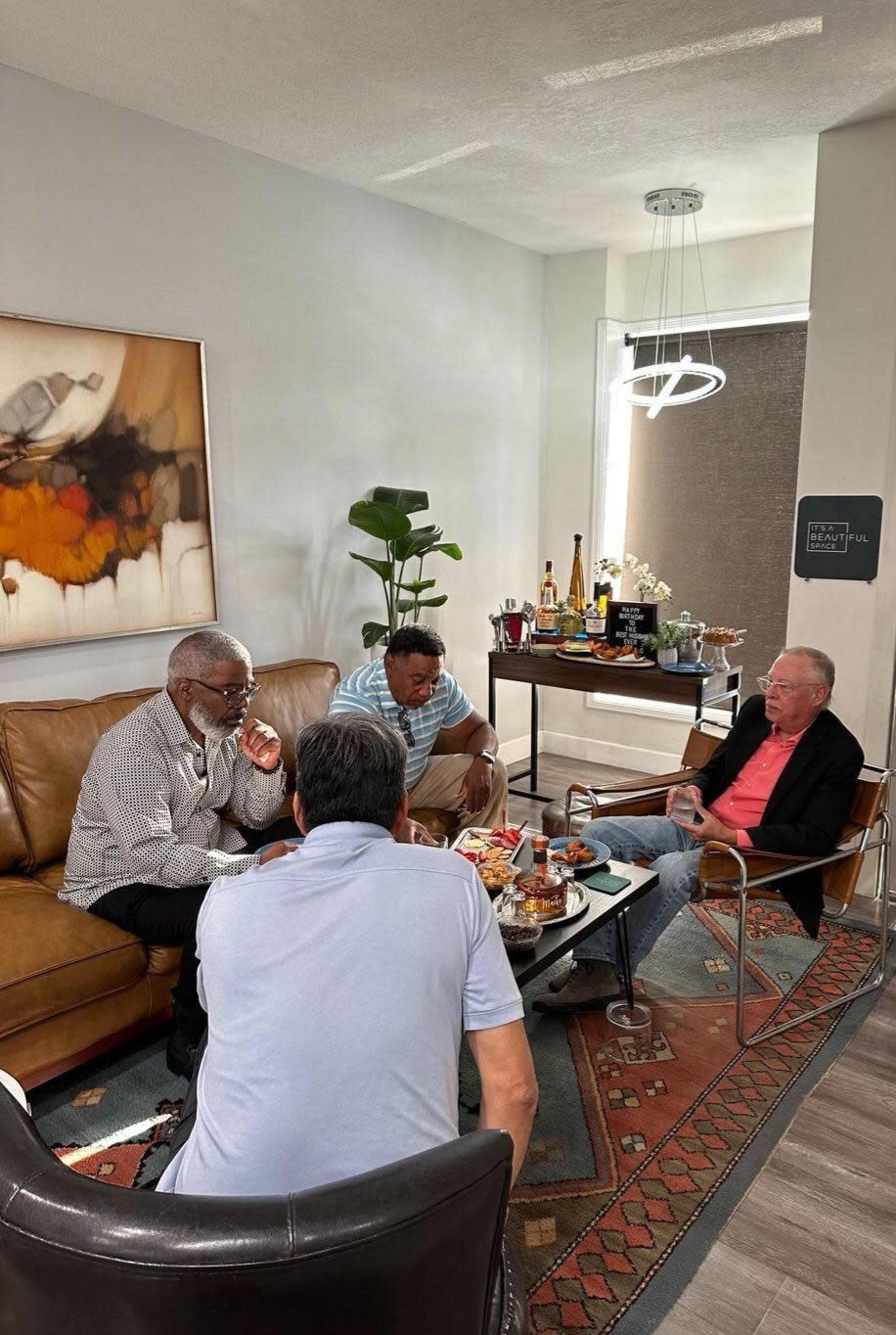 Four men are seated in a modern living space, engaged in conversation while surrounded by food and drinks on a coffee table.
