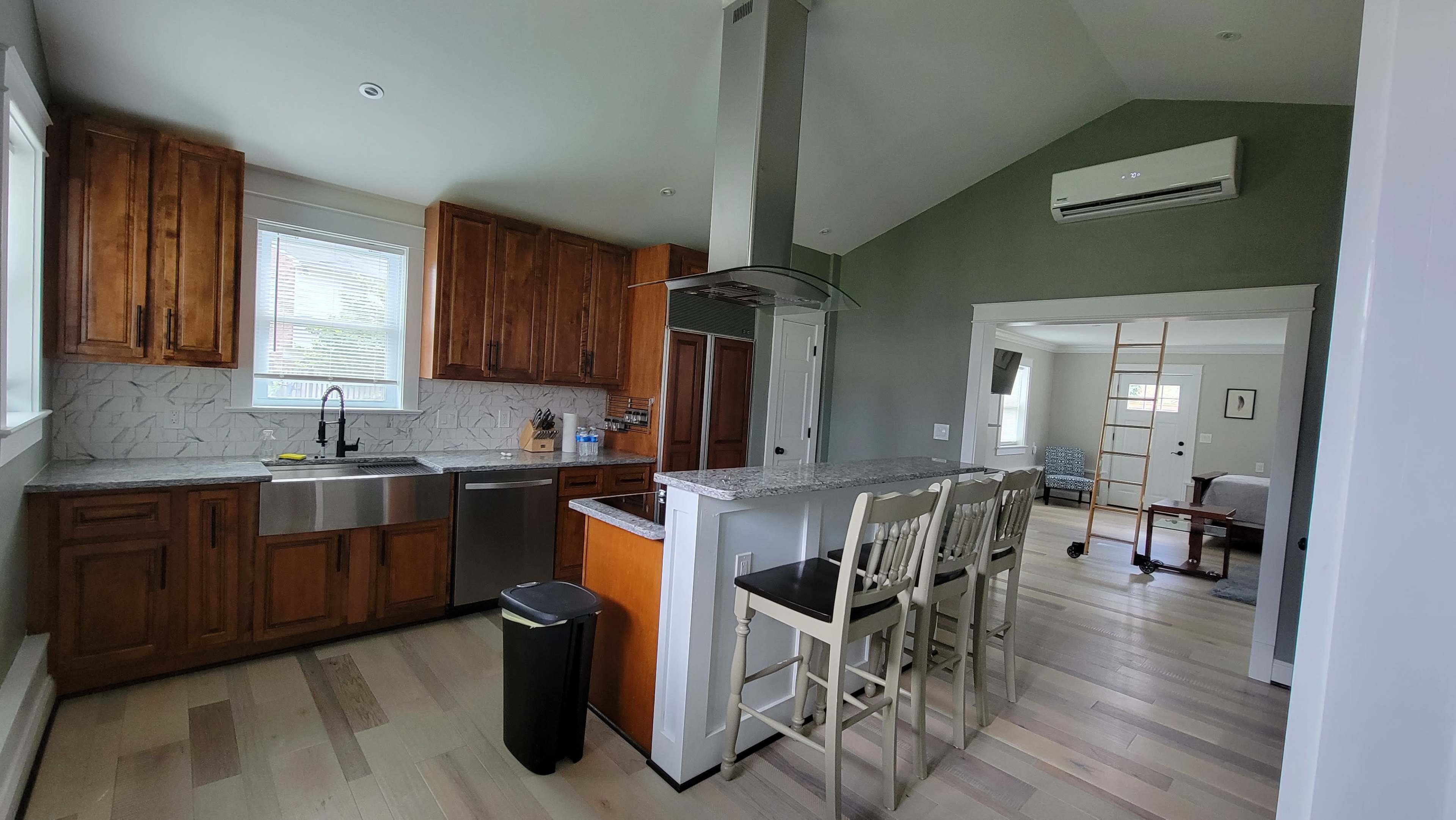 The image shows a modern kitchen with wooden cabinets, a stainless steel dishwasher and refrigerator, and a breakfast bar with four chairs.