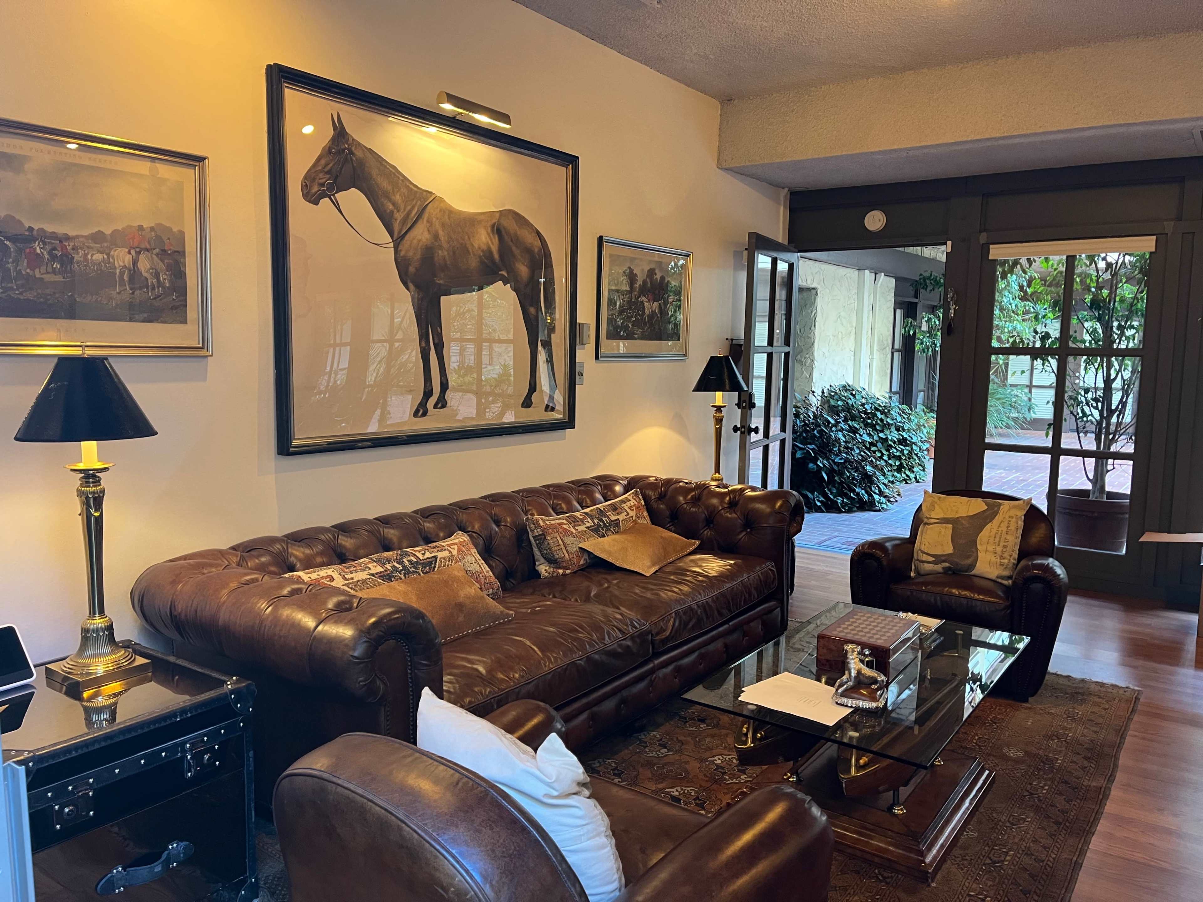 A living room with a brown leather sofa and chairs, a glass coffee table, and a large painting of a horse on the wall.