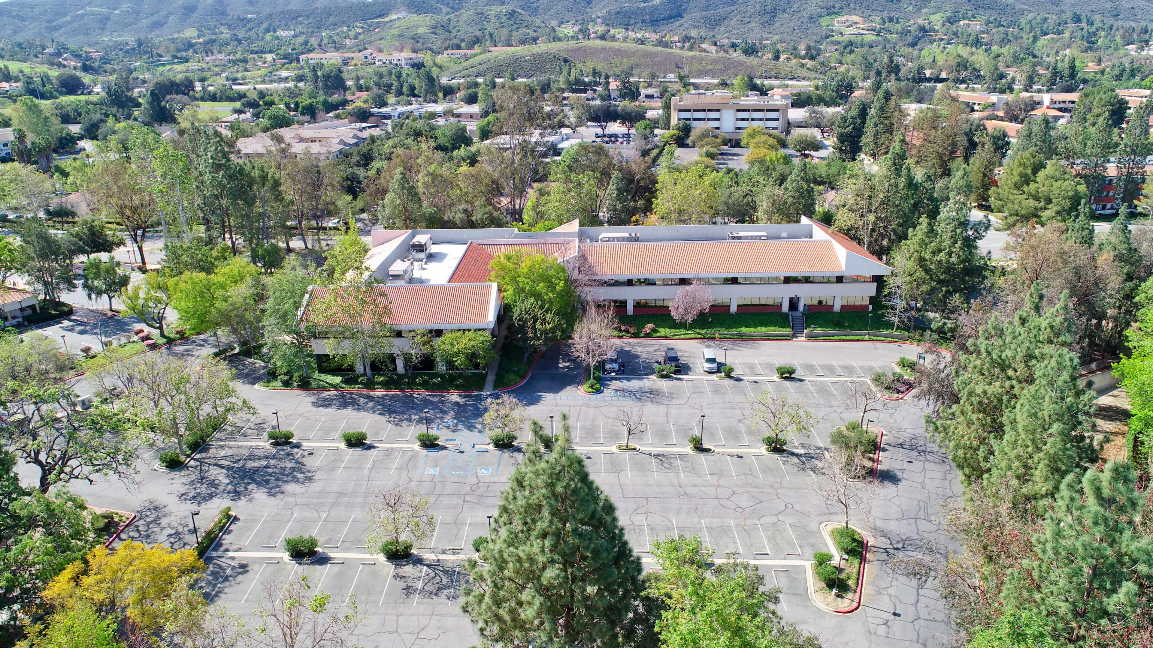 An aerial view shows a large, empty parking lot in front of a commercial building surrounded by trees and hills.