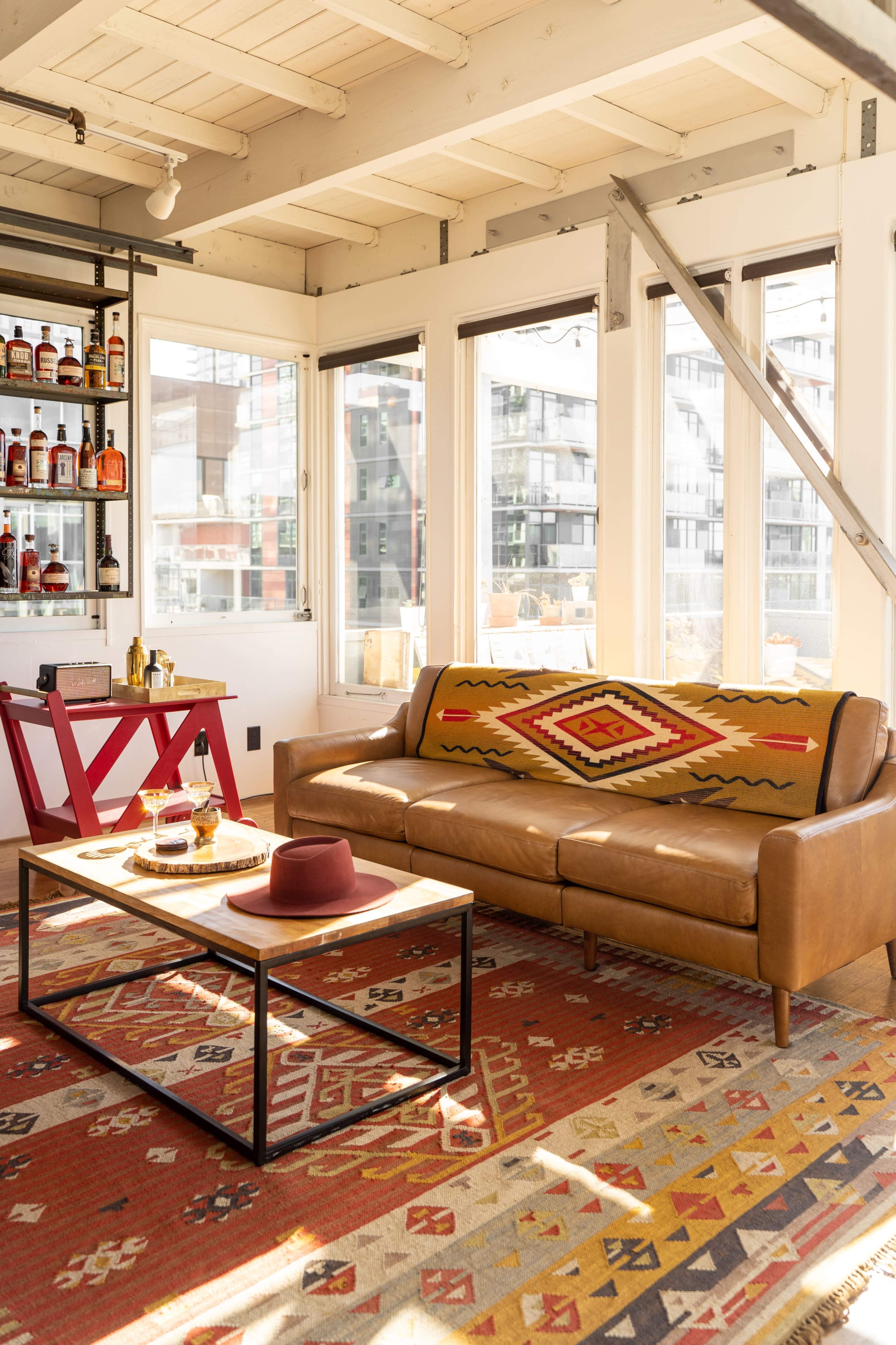 A leather sofa with a patterned blanket sits in a bright room featuring large windows, a wooden ceiling, and a coffee table with a hat and decorative tray.