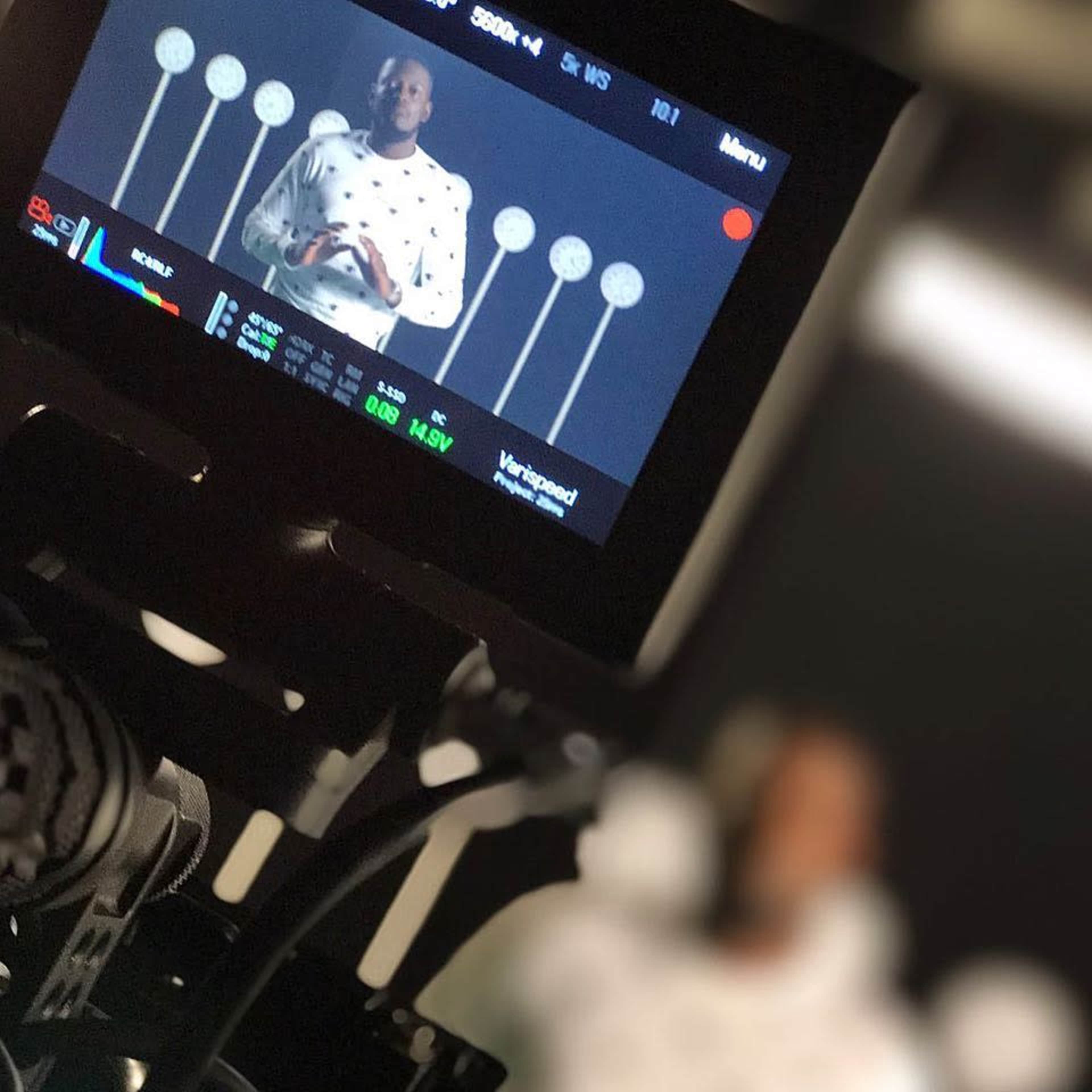 A person wearing a patterned sweatshirt is speaking in front of several lighted circles, viewed through a camera monitor.