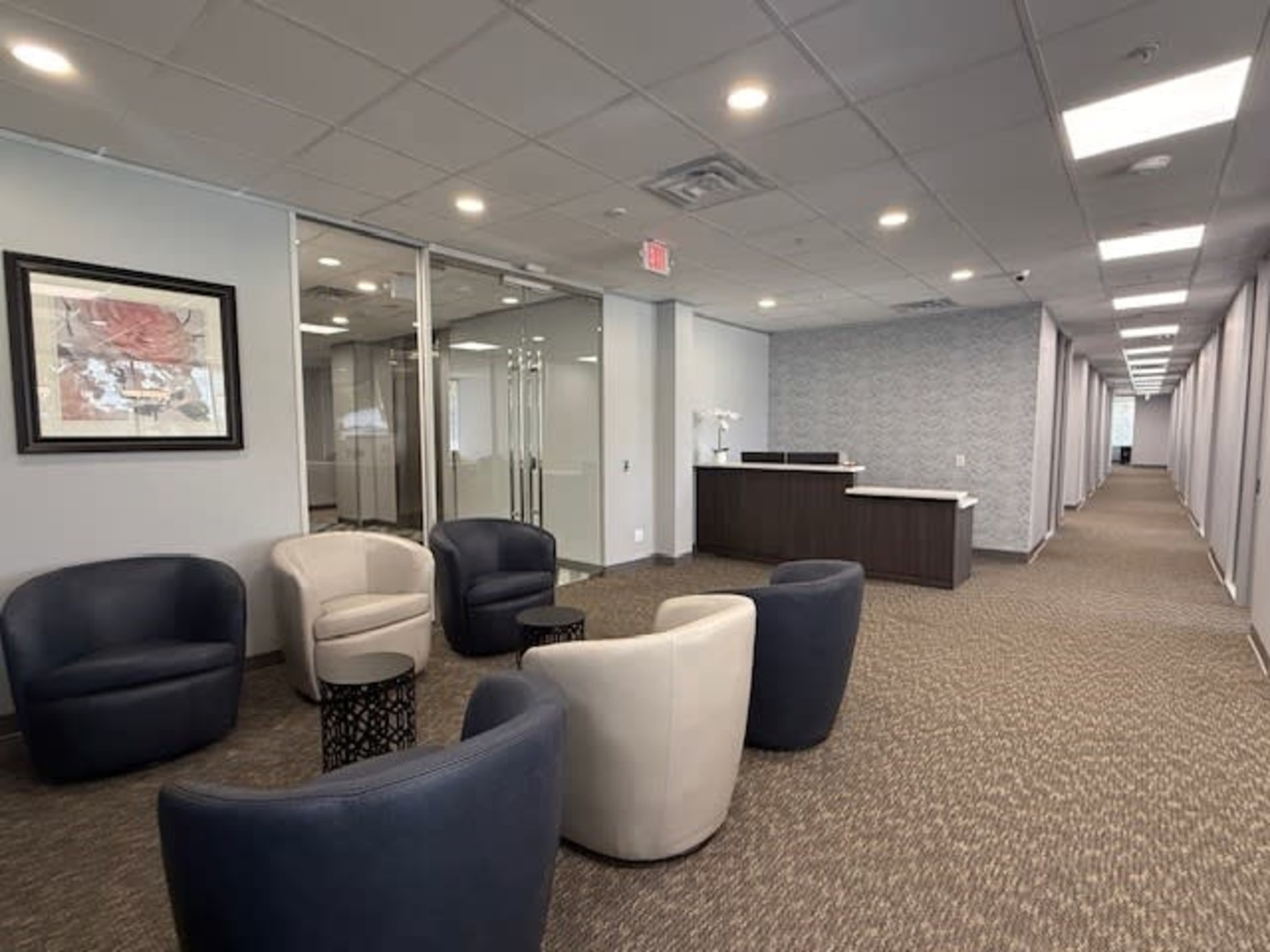 The image shows a modern office reception area with a seating arrangement of upholstered chairs, a front desk, and a long corridor in the background.