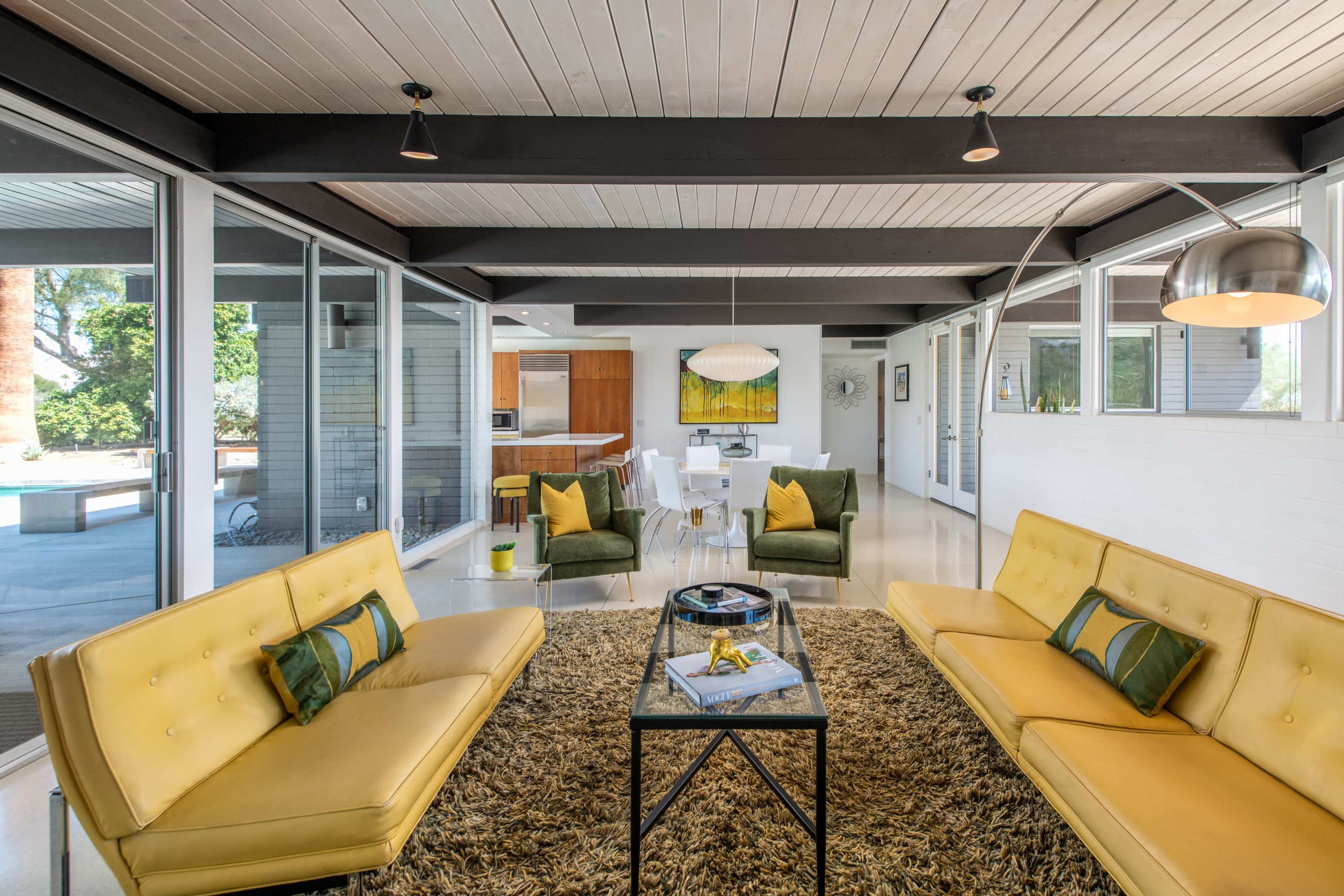 A modern living room with two yellow sofas facing a glass coffee table, surrounded by green armchairs and large windows letting in natural light.