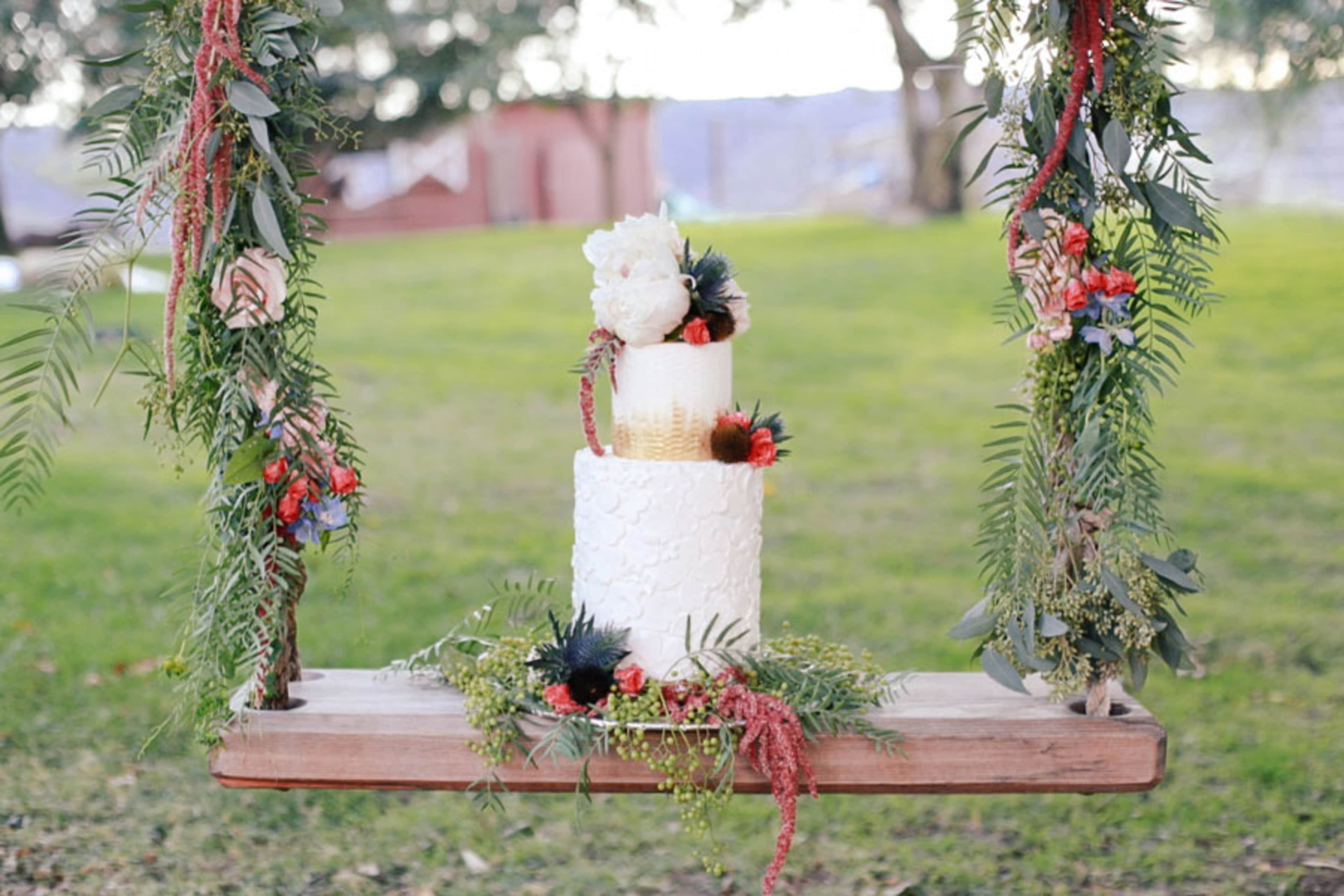A two-tiered wedding cake adorned with flowers and greenery is displayed on a wooden swing in a grassy outdoor setting.