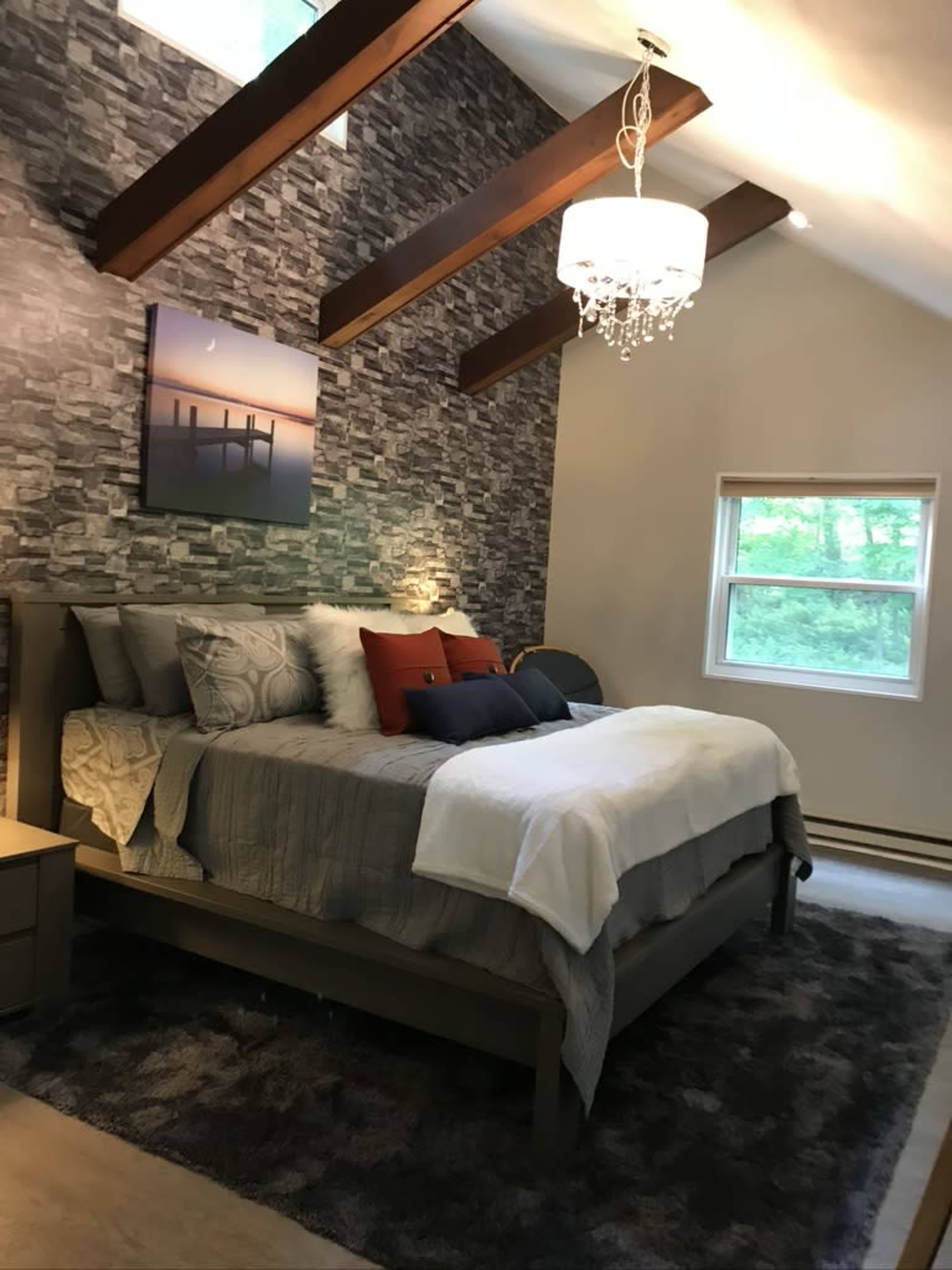 A bedroom with a large bed, a chandelier, and stone accent walls, illuminated by natural light from a nearby window.