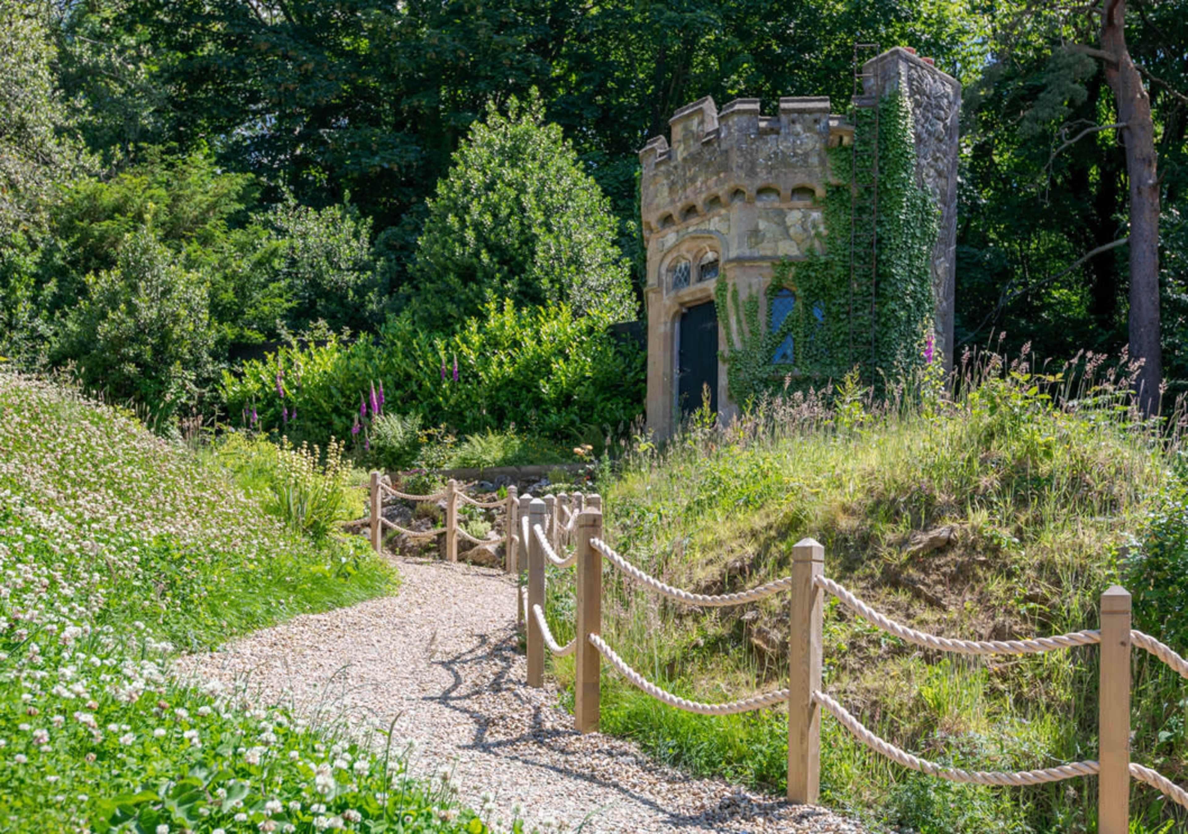 A gravel path lined with a wooden fence leads to a small, round stone structure covered in greenery.