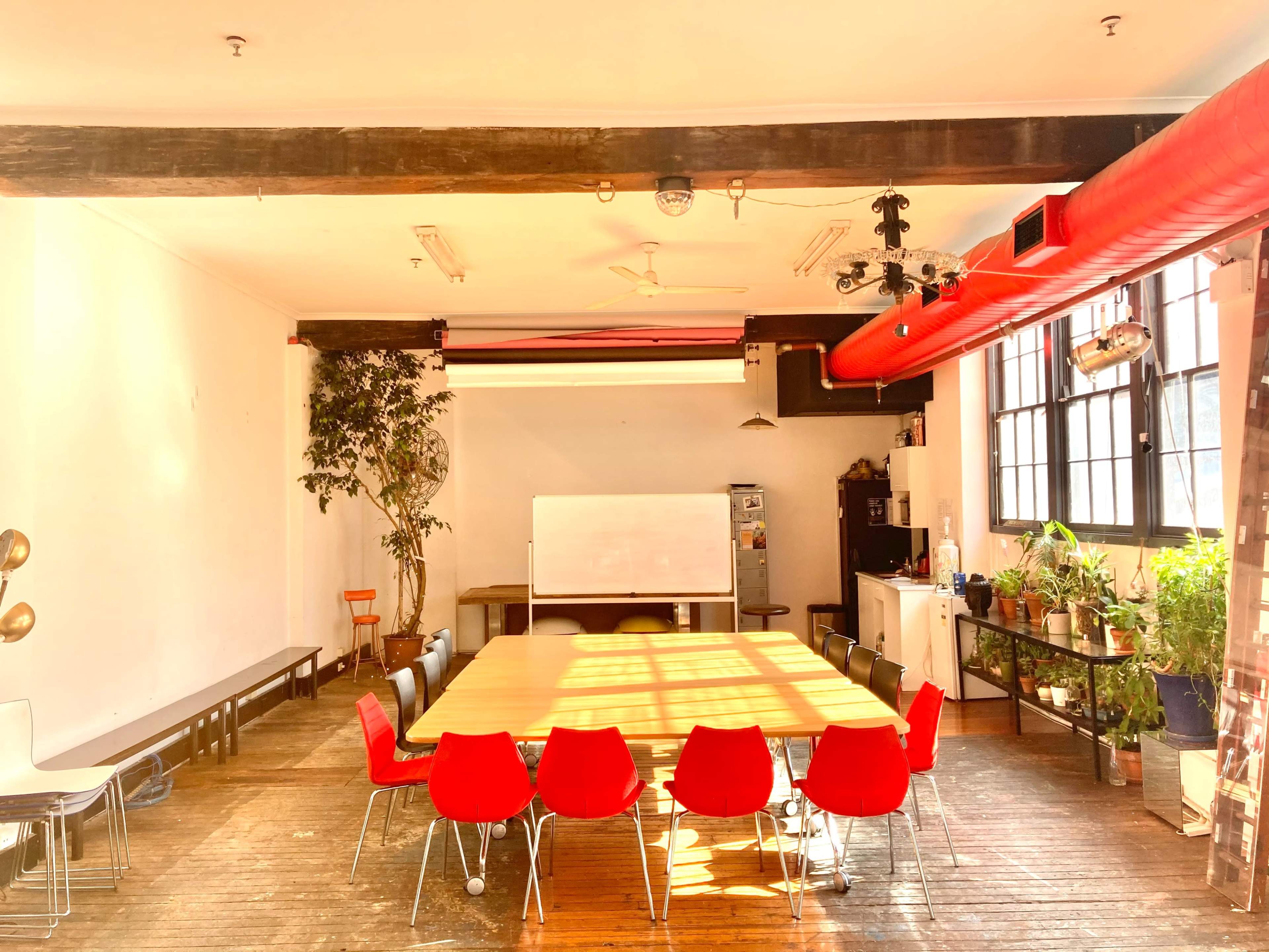 A large meeting room features a wooden table surrounded by red and white chairs, with plants and a whiteboard against the walls.