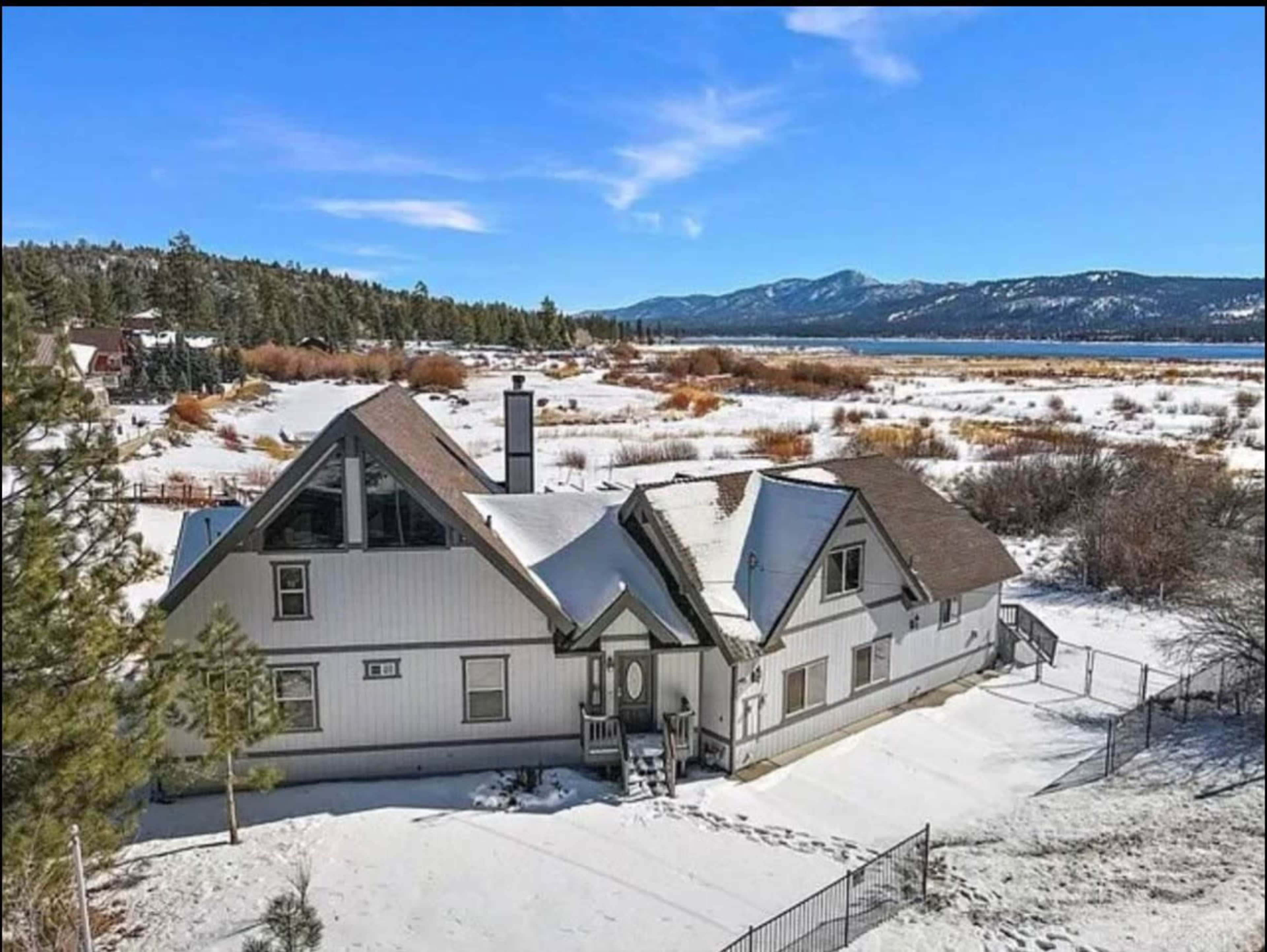 A large, multi-level house with a steep roof sits in a snowy landscape, surrounded by trees and mountains in the background.