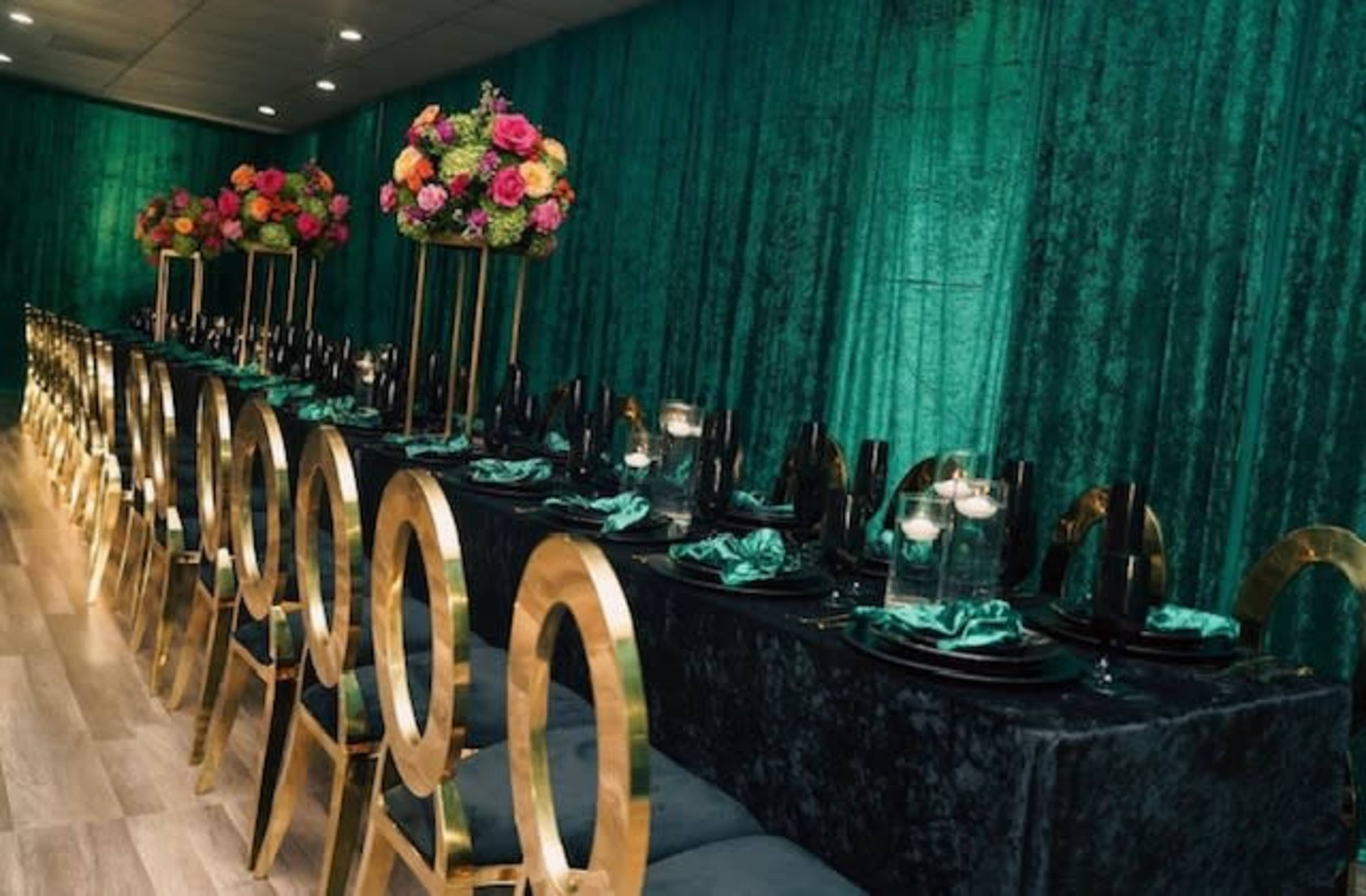 A long dining table is set with black tableware and surrounded by gold chairs, decorated with colorful floral centerpieces against a green backdrop.