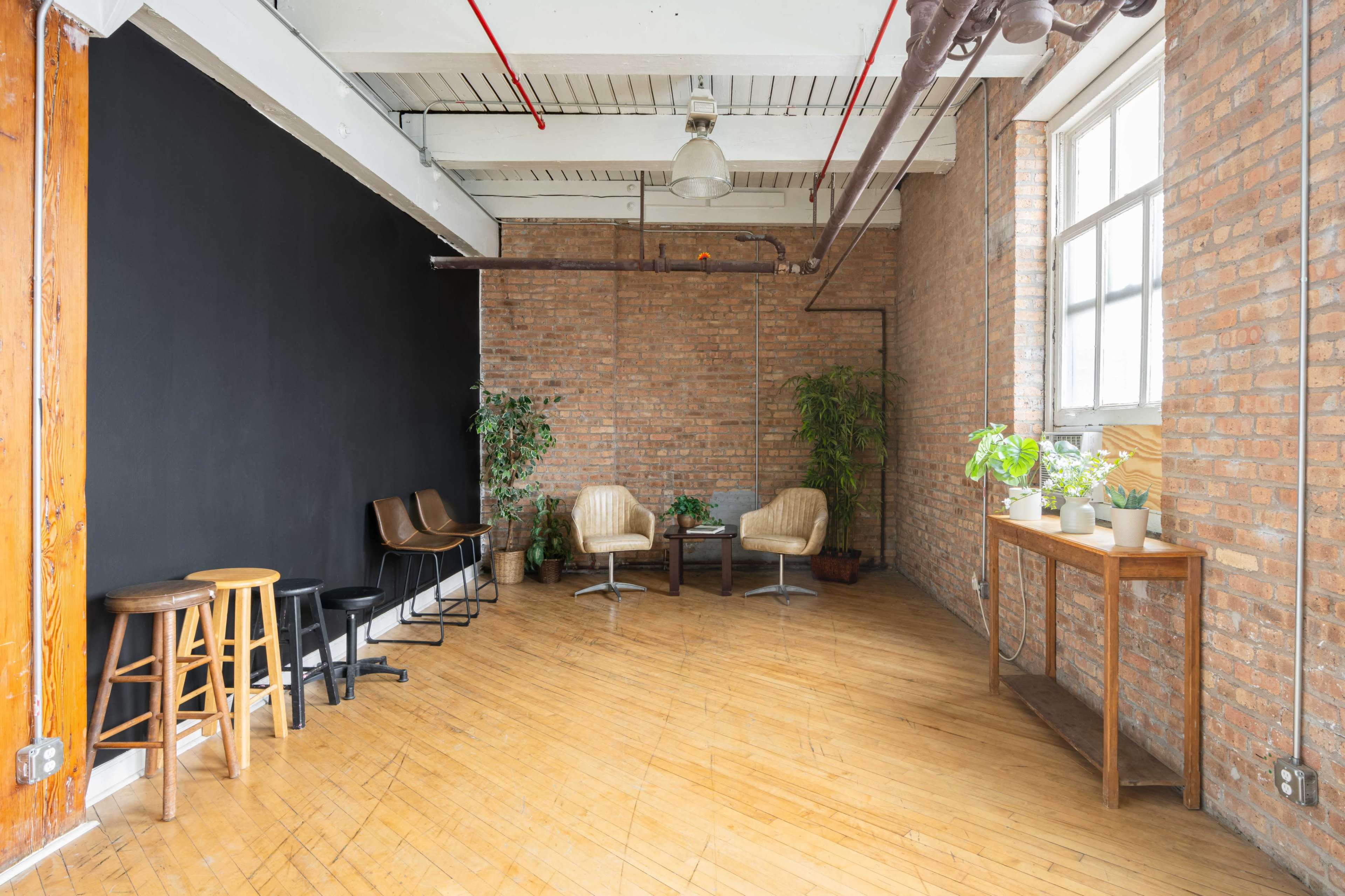 The interior of a spacious room features a black wall, wooden flooring, and seating arrangements, including stools and chairs, along with several potted plants.