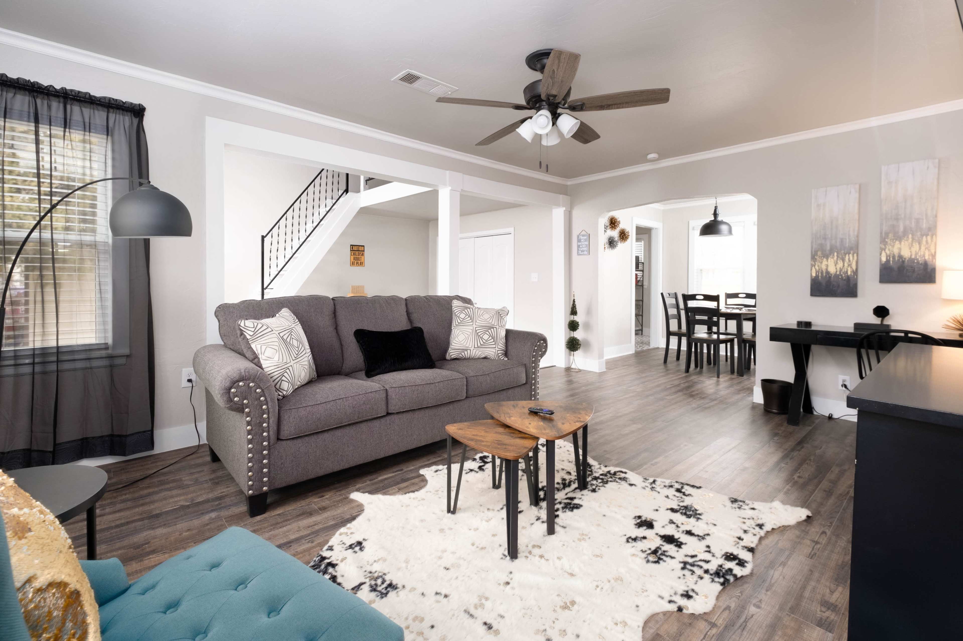 The image showcases a modern living room with a gray sofa, a coffee table set on a patterned rug, and an open layout leading to a dining area.