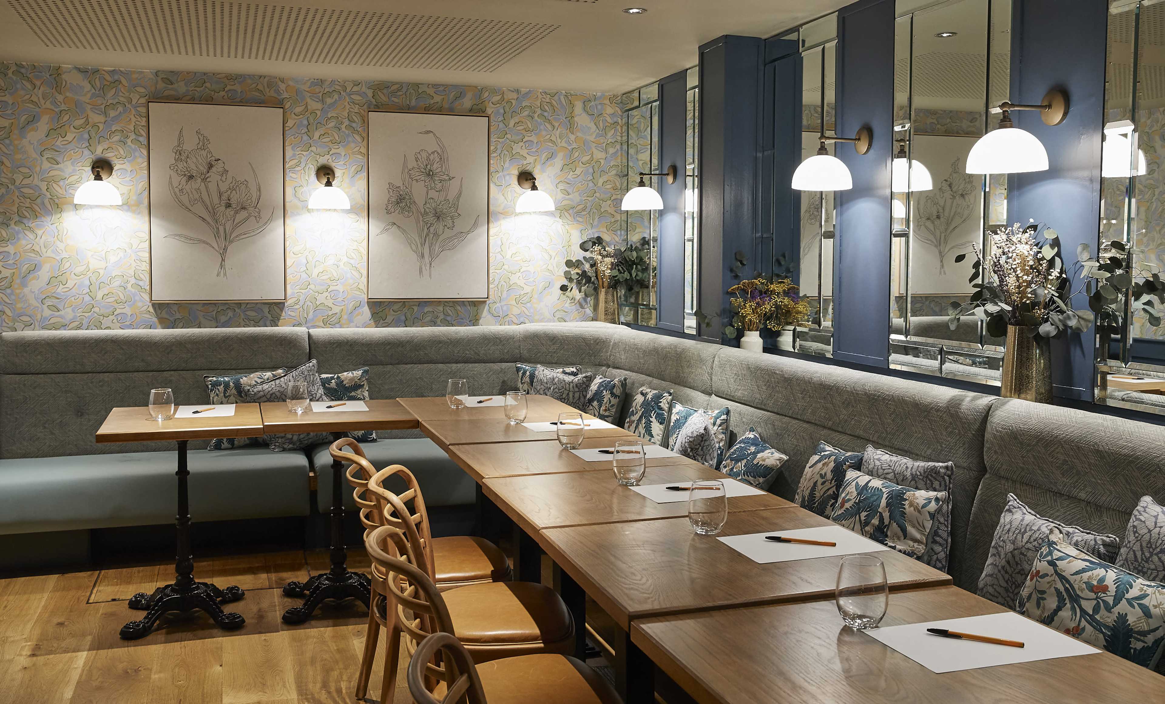The image shows a modern restaurant interior featuring a long wooden table with stationery, surrounded by upholstered seating and decorative floral wallpaper.