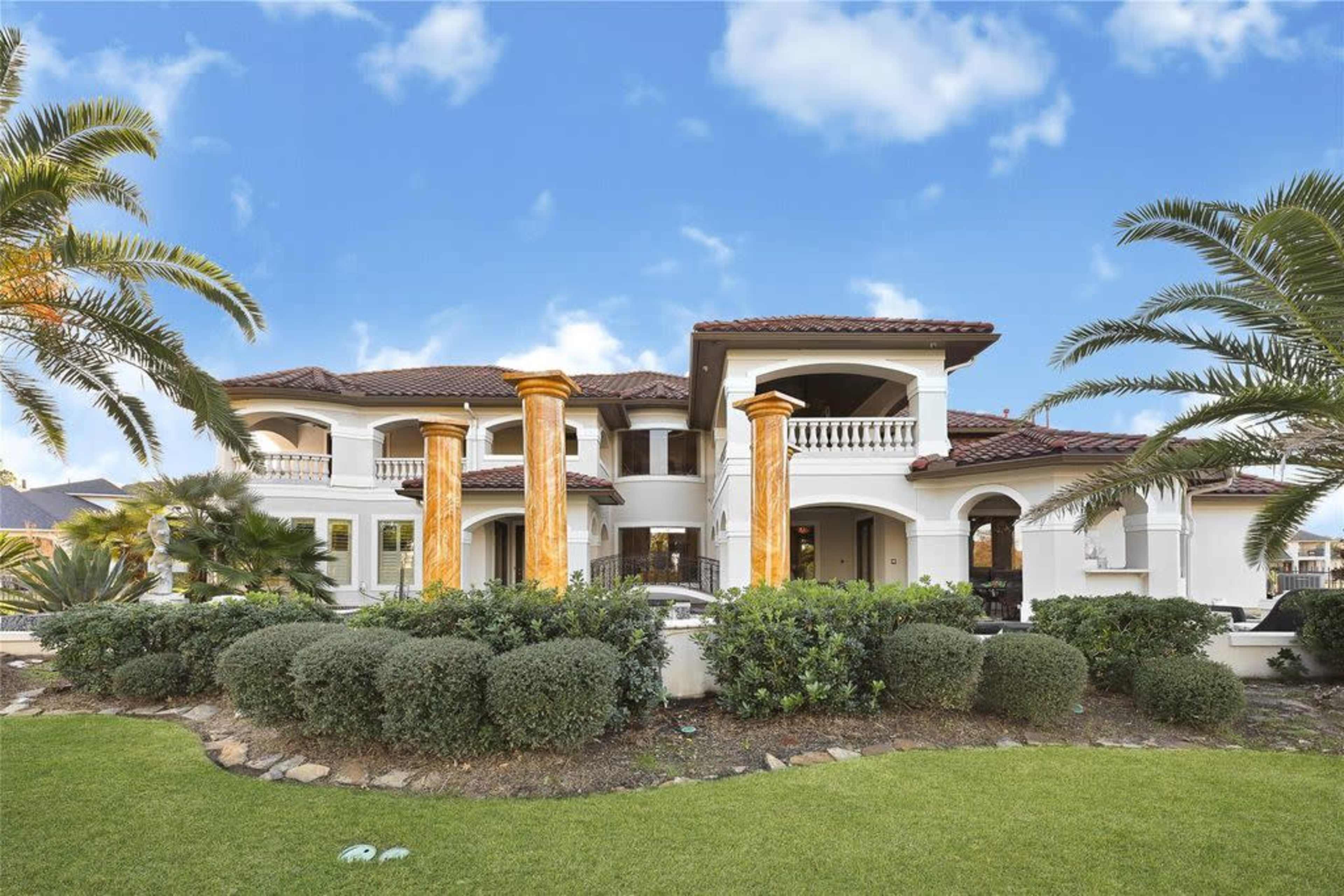 The image depicts a large, two-story Mediterranean-style house with stone columns, surrounded by palm trees and a well-maintained lawn.