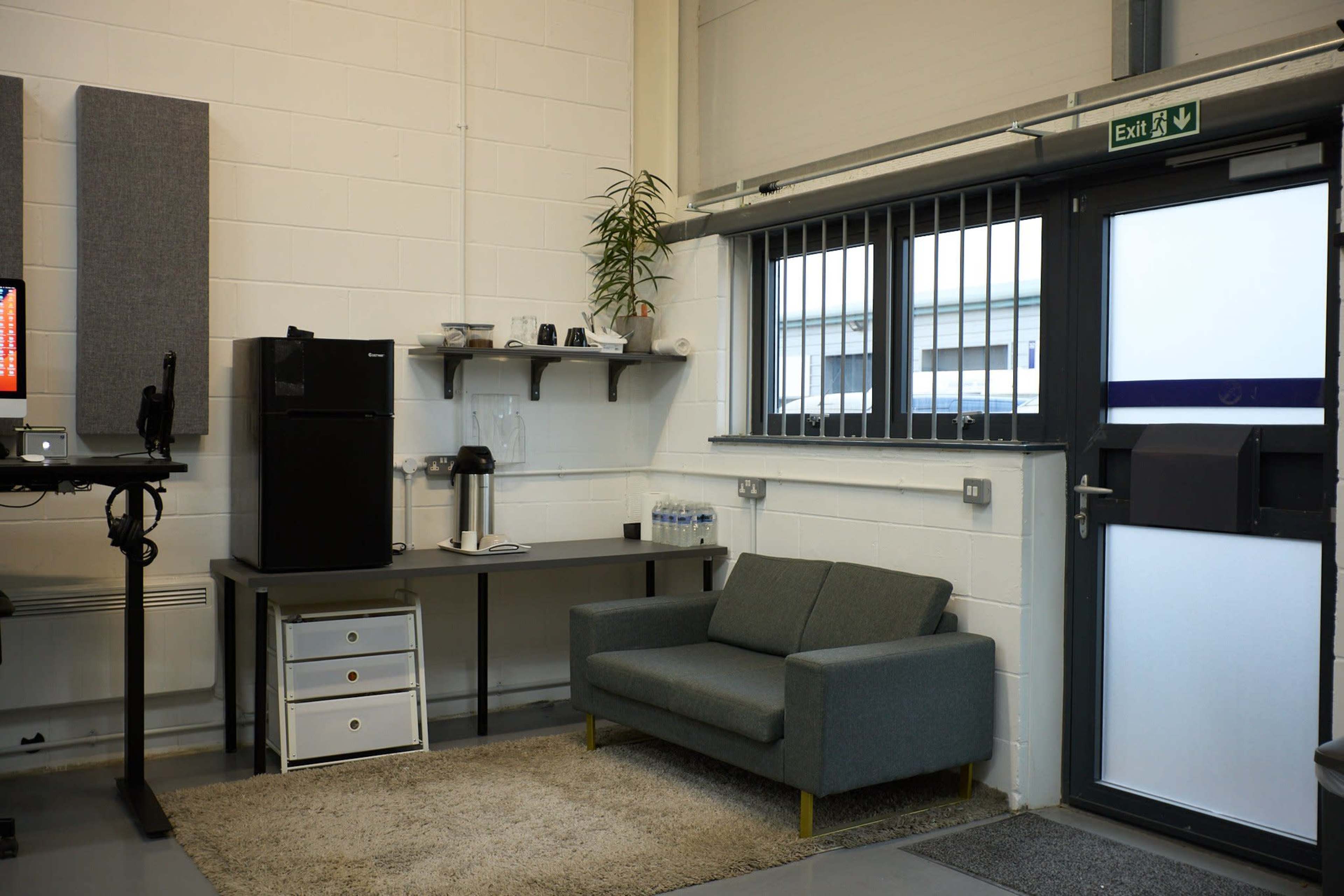A modern office space featuring a small gray couch, a desk with a black refrigerator, and a window with security bars.