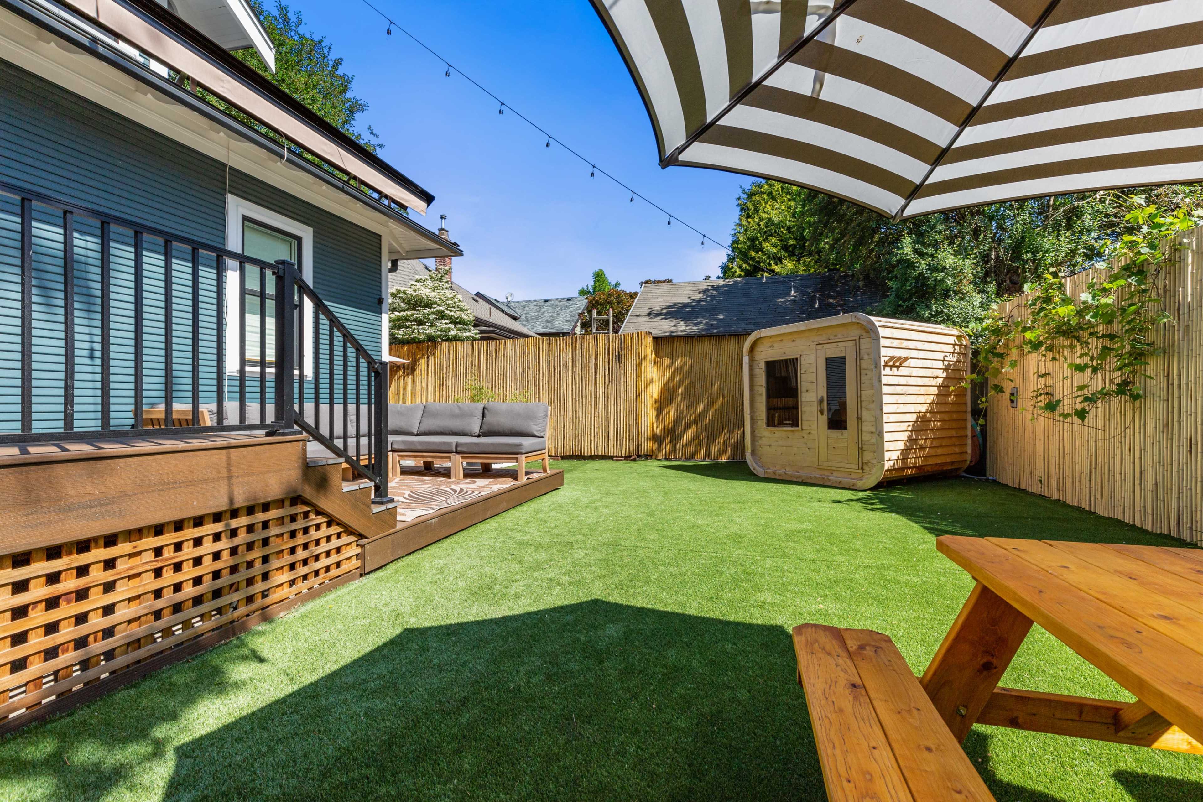 A backyard with a green artificial lawn, a wooden picnic table, a lounge area with gray seating, and a small wooden structure against a wooden fence.