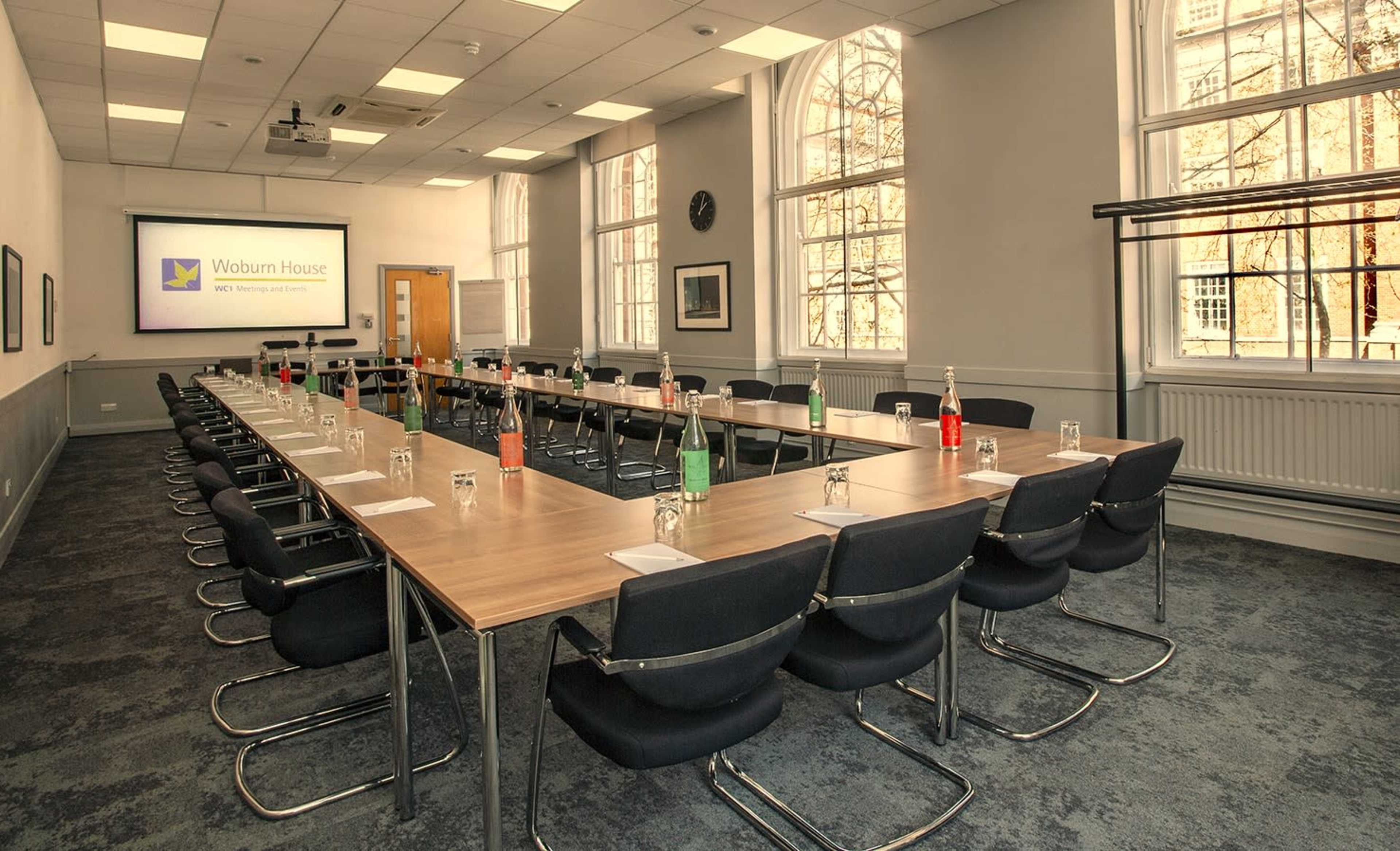 A meeting room is set up with rectangular tables arranged in a U-shape, featuring chairs and various drinks on the tables, with a presentation screen at one end.