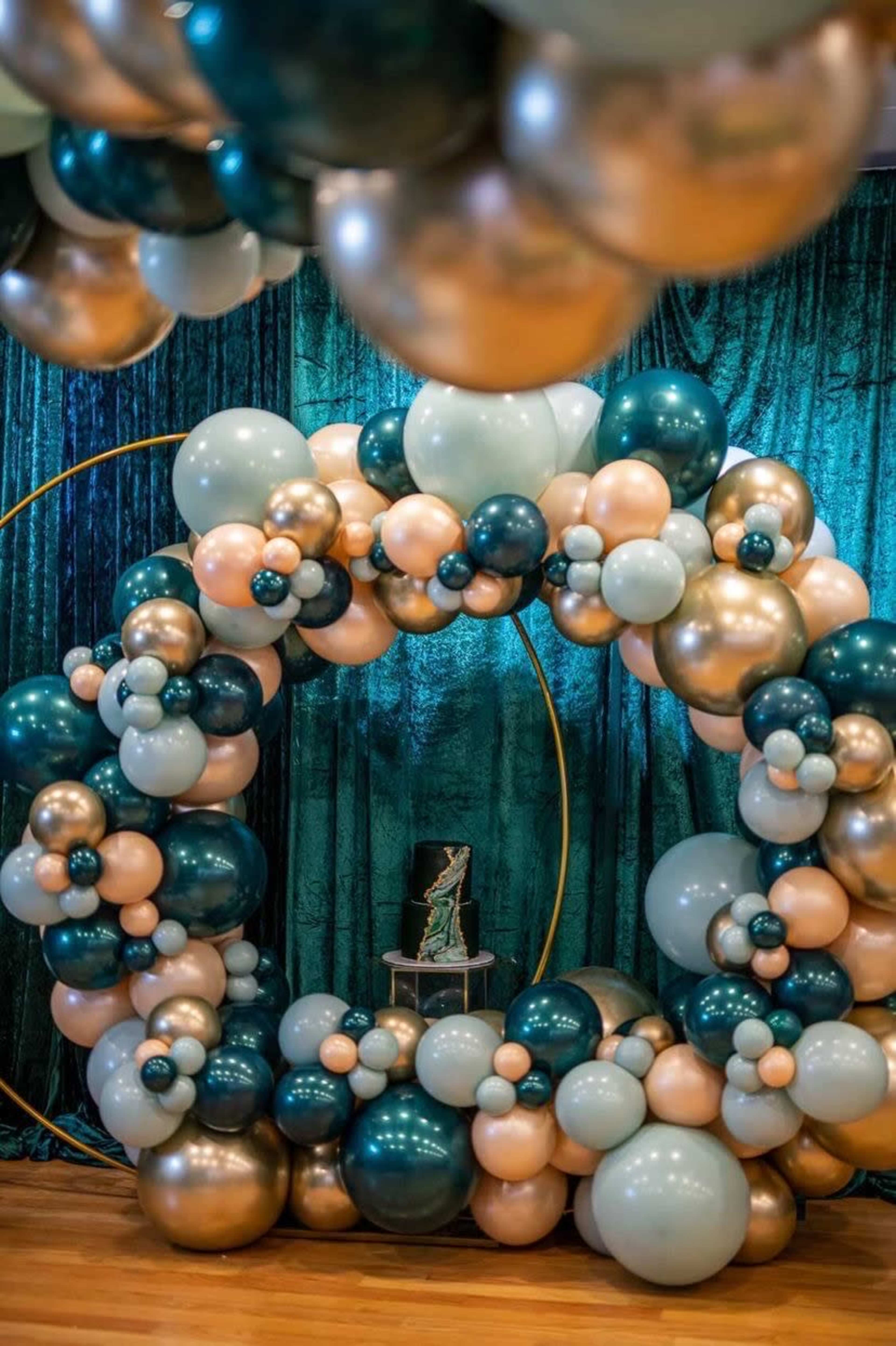 A decorative balloon arch in shades of teal, peach, and gold, set against a shimmering green backdrop.