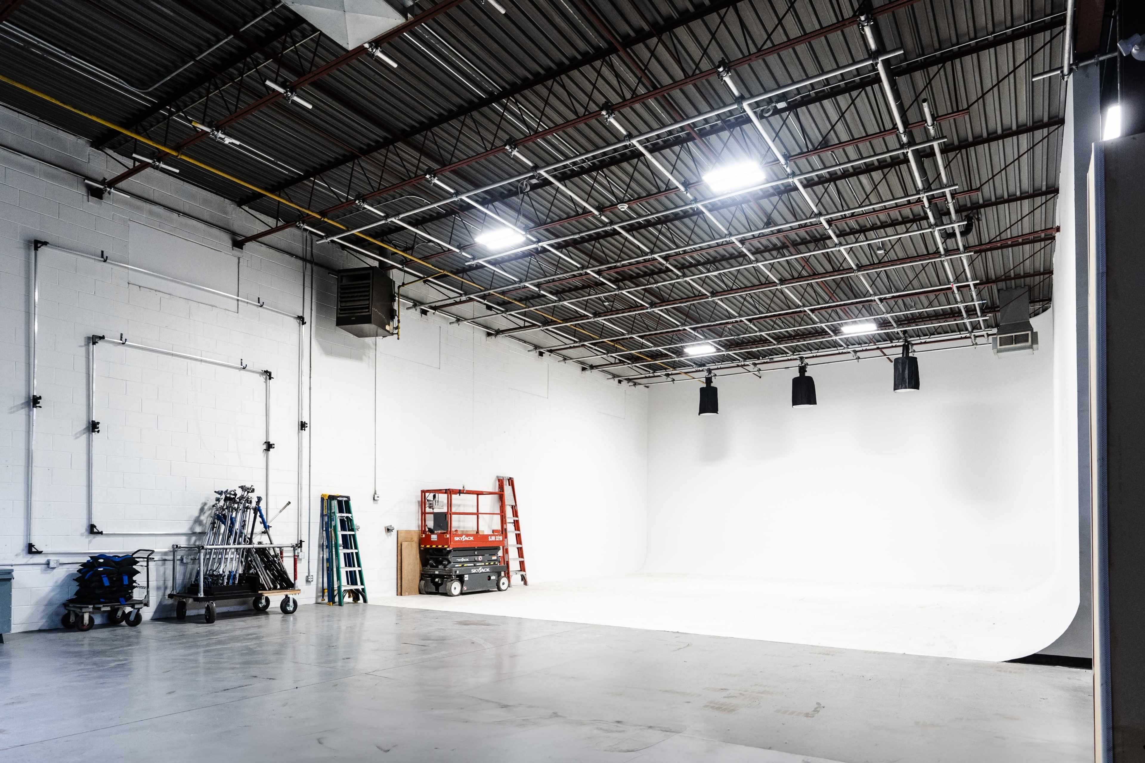An empty photography studio features a large white backdrop, several ladders, and various lighting equipment along the walls.