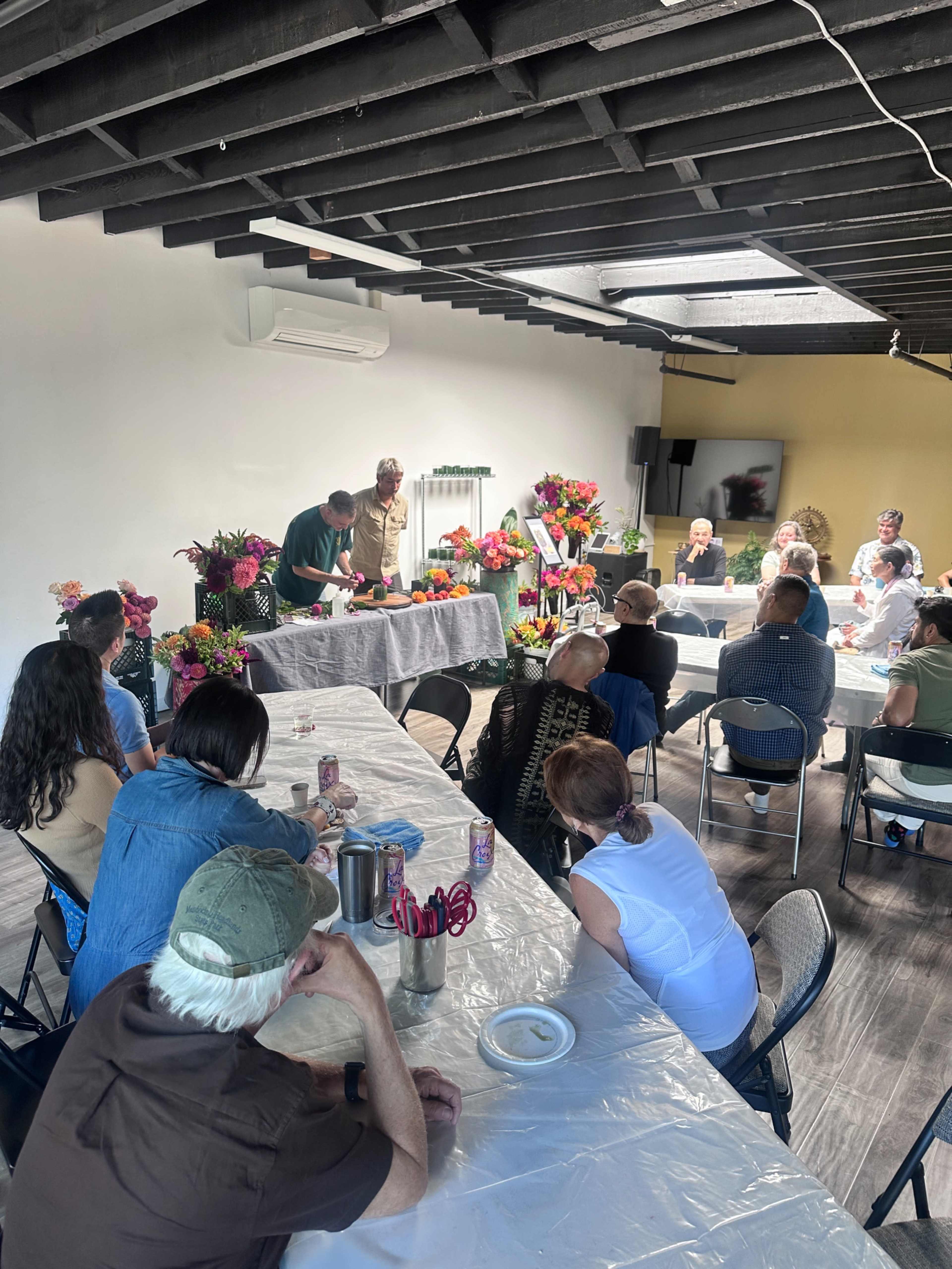 A gathering of people sits around tables filled with flowers while a presenter demonstrates a technique at the front of the room.