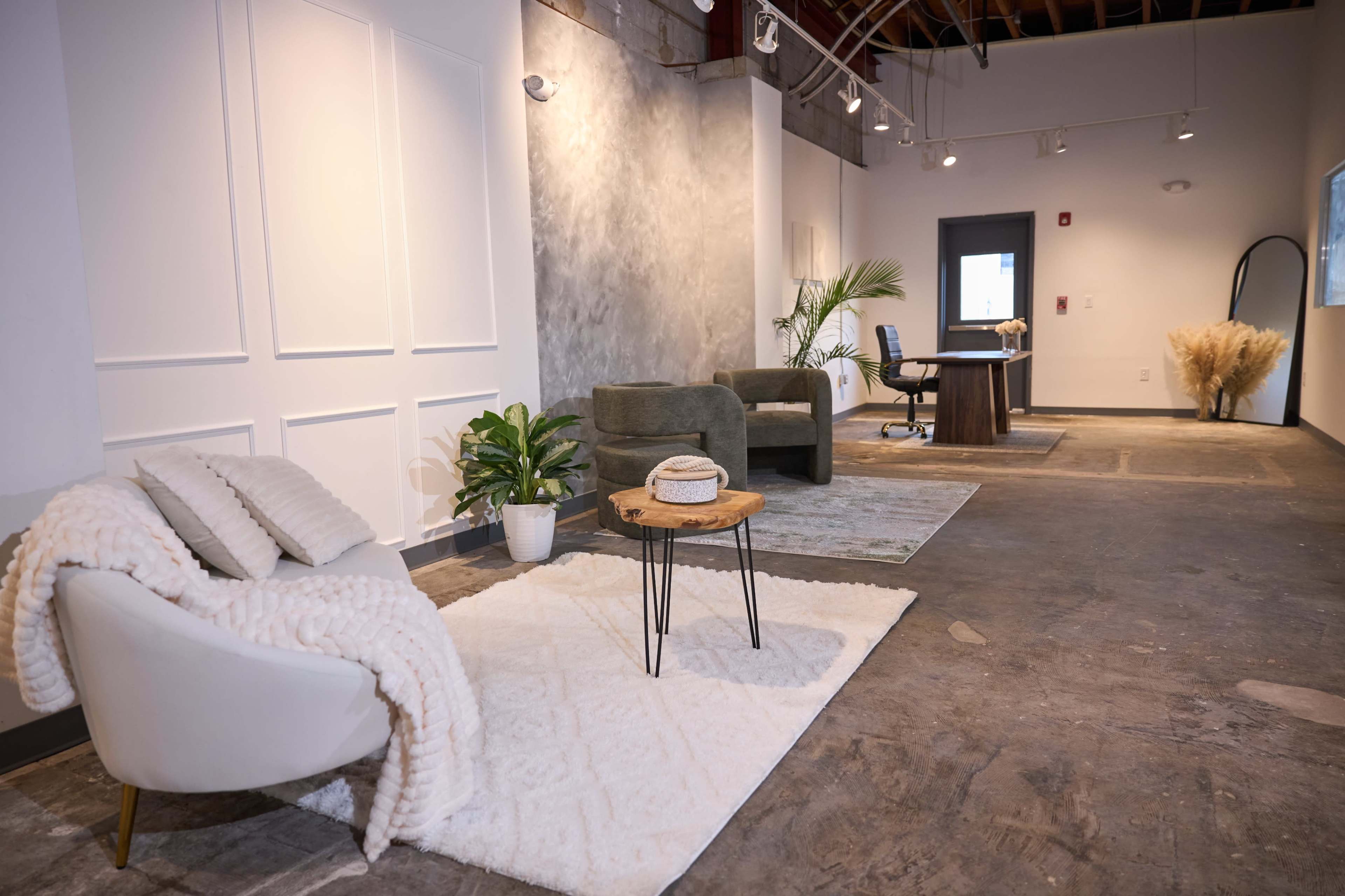 The image shows a spacious interior with modern furniture, including a seating area with a white armchair and a small wooden table, along with a desk and decor elements like plants and rugs.