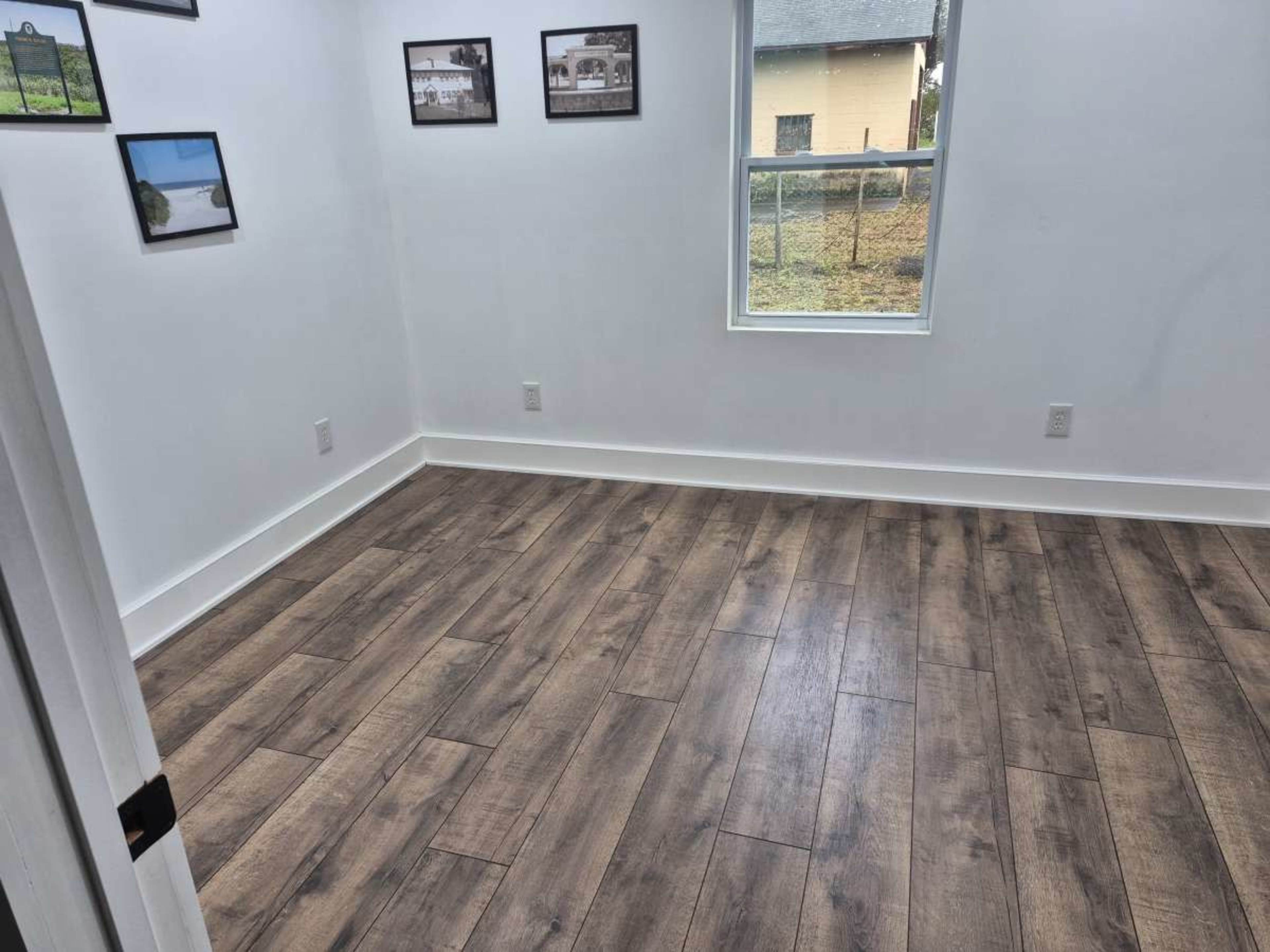 The image shows a vacant room with wooden floors, white walls, and several framed pictures on the wall.