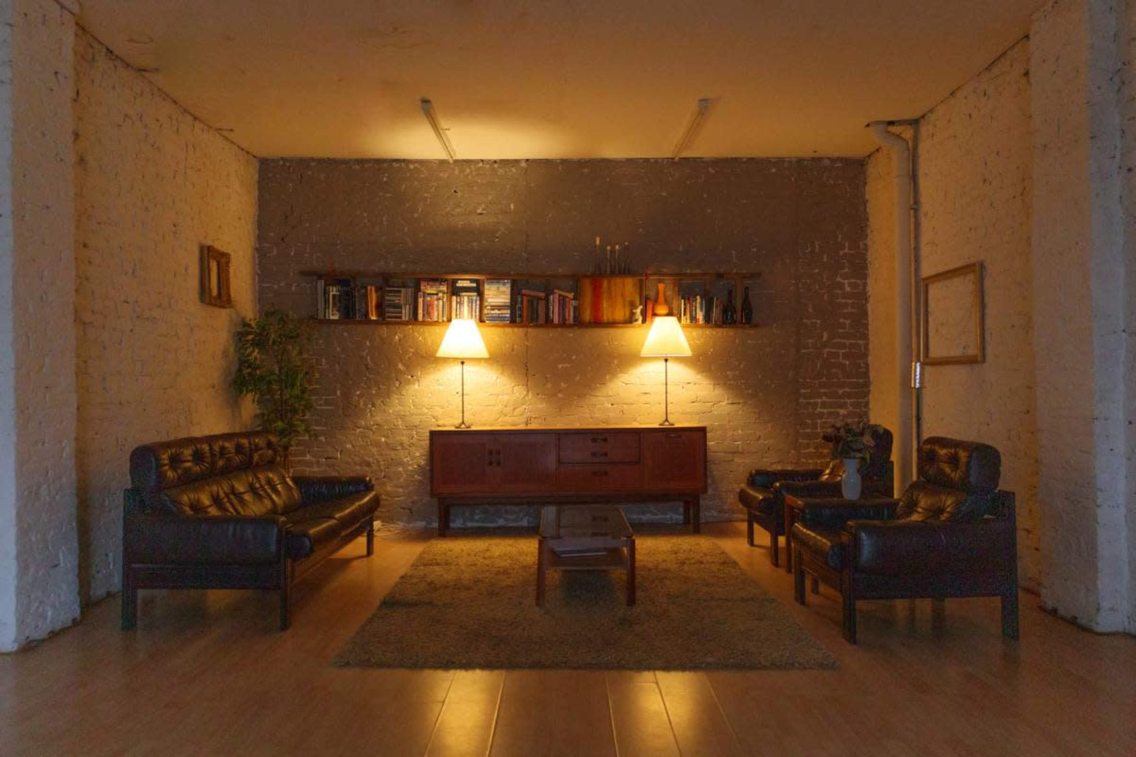 The image shows a minimalist living room with two dark leather sofas and two matching armchairs arranged around a wooden coffee table, illuminated by two lamps on a shelf above a wooden console.