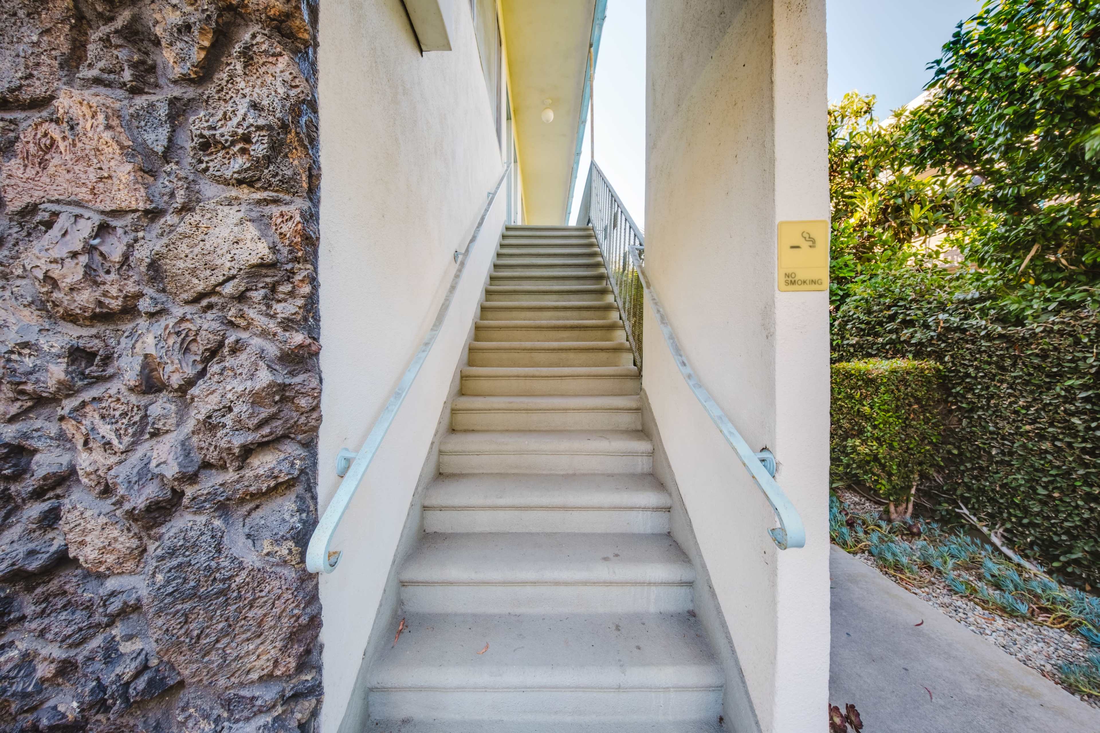 A set of concrete stairs leads upward between two walls, one made of stone and the other smooth, with greenery visible on either side.