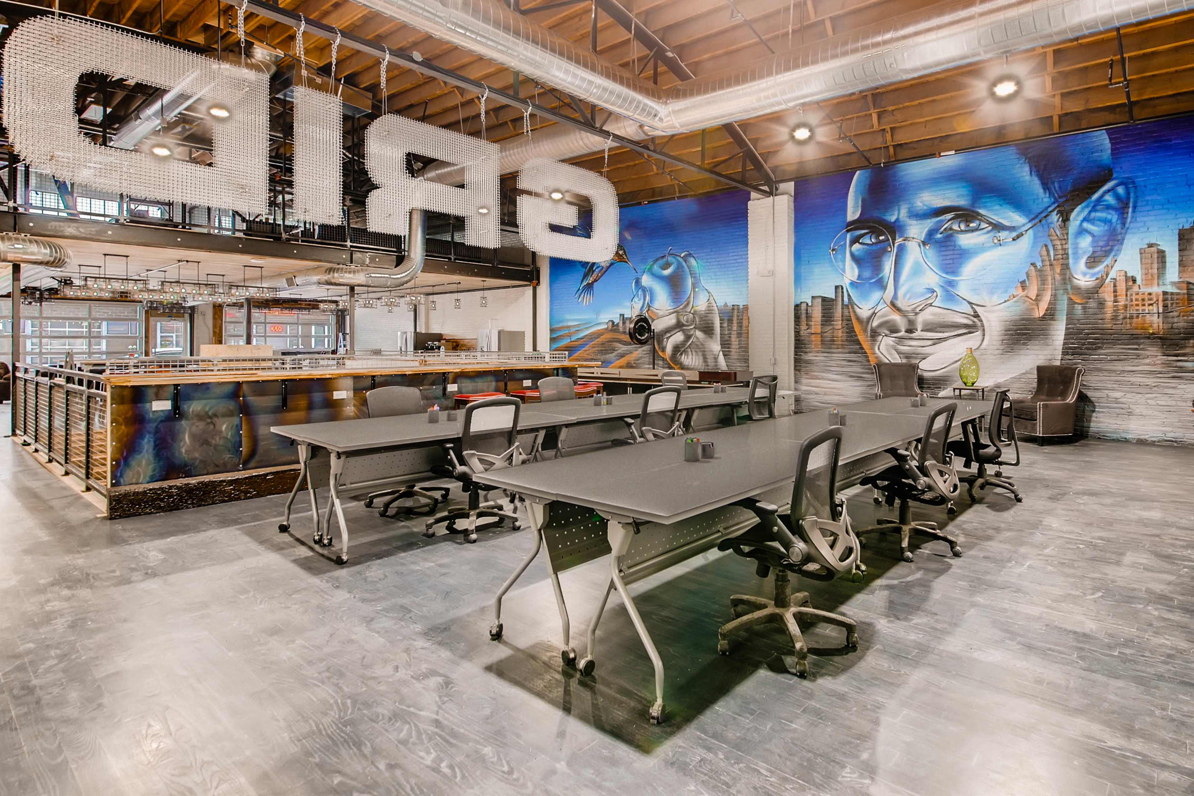 The image shows a modern coworking space with several black desks and a large mural of a person in glasses on the wall.