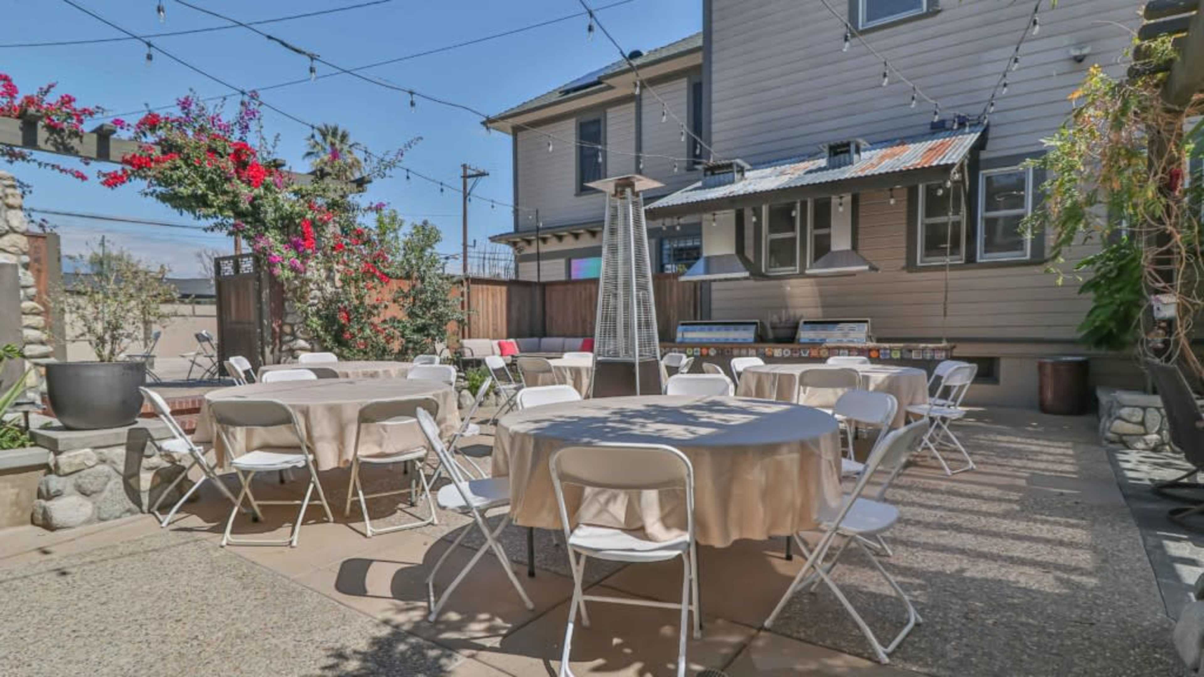 The image shows a spacious outdoor patio area with several round tables surrounded by string lights and potted plants.