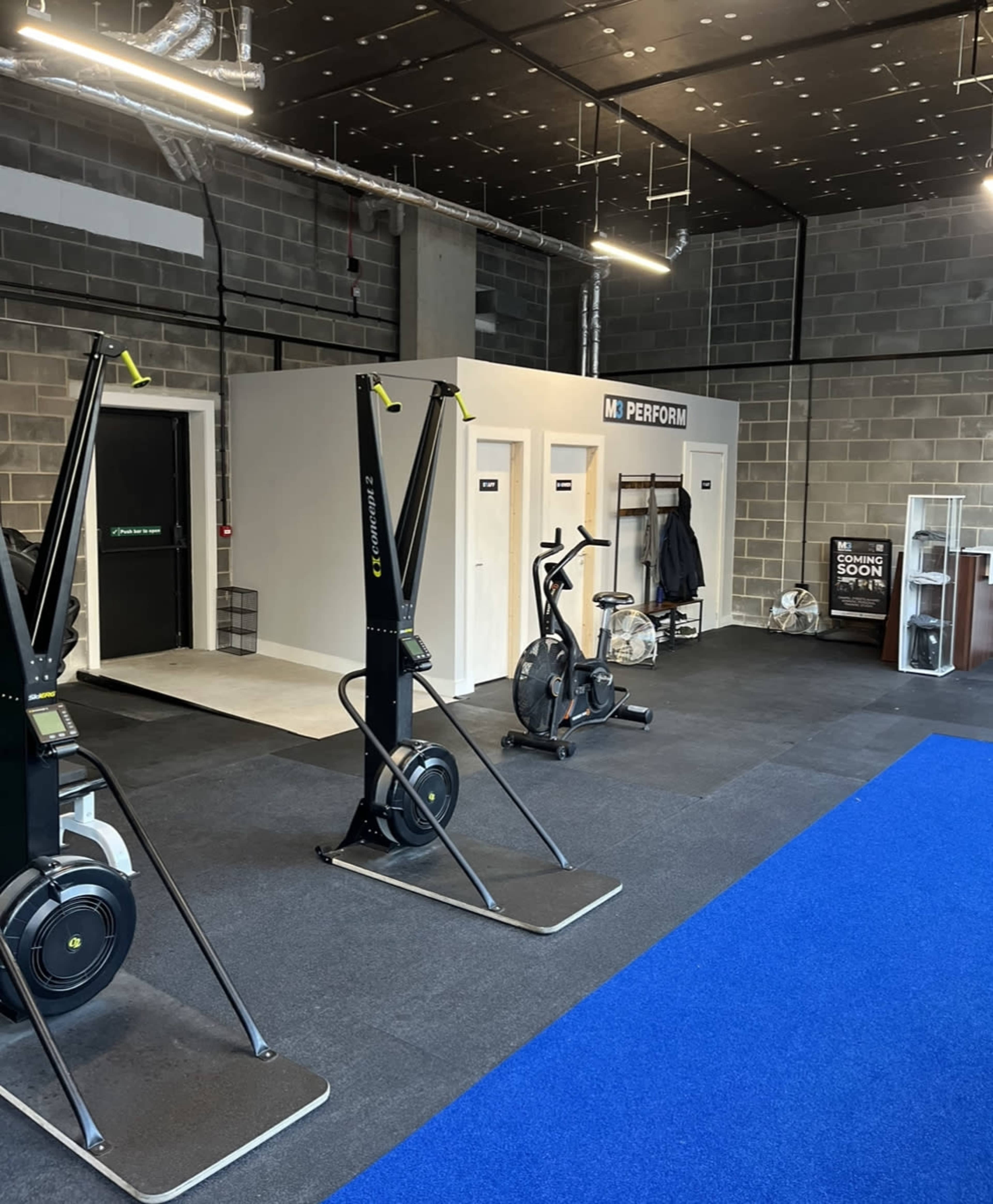 A modern gym interior with exercise equipment, including two ski ergs and an elliptical, situated on a blue carpeted floor.