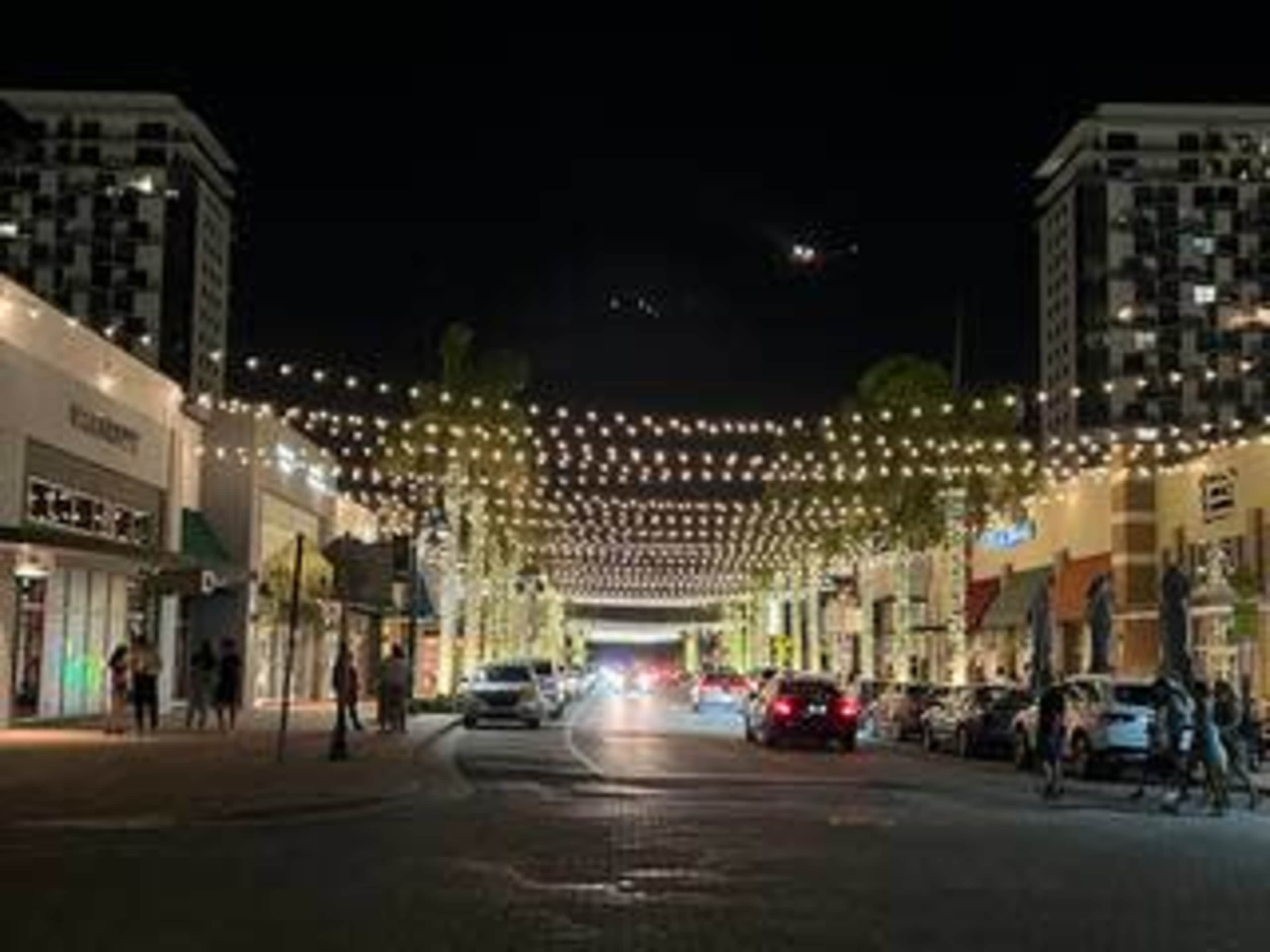The scene shows a well-lit street lined with shops and adorned with string lights at night.