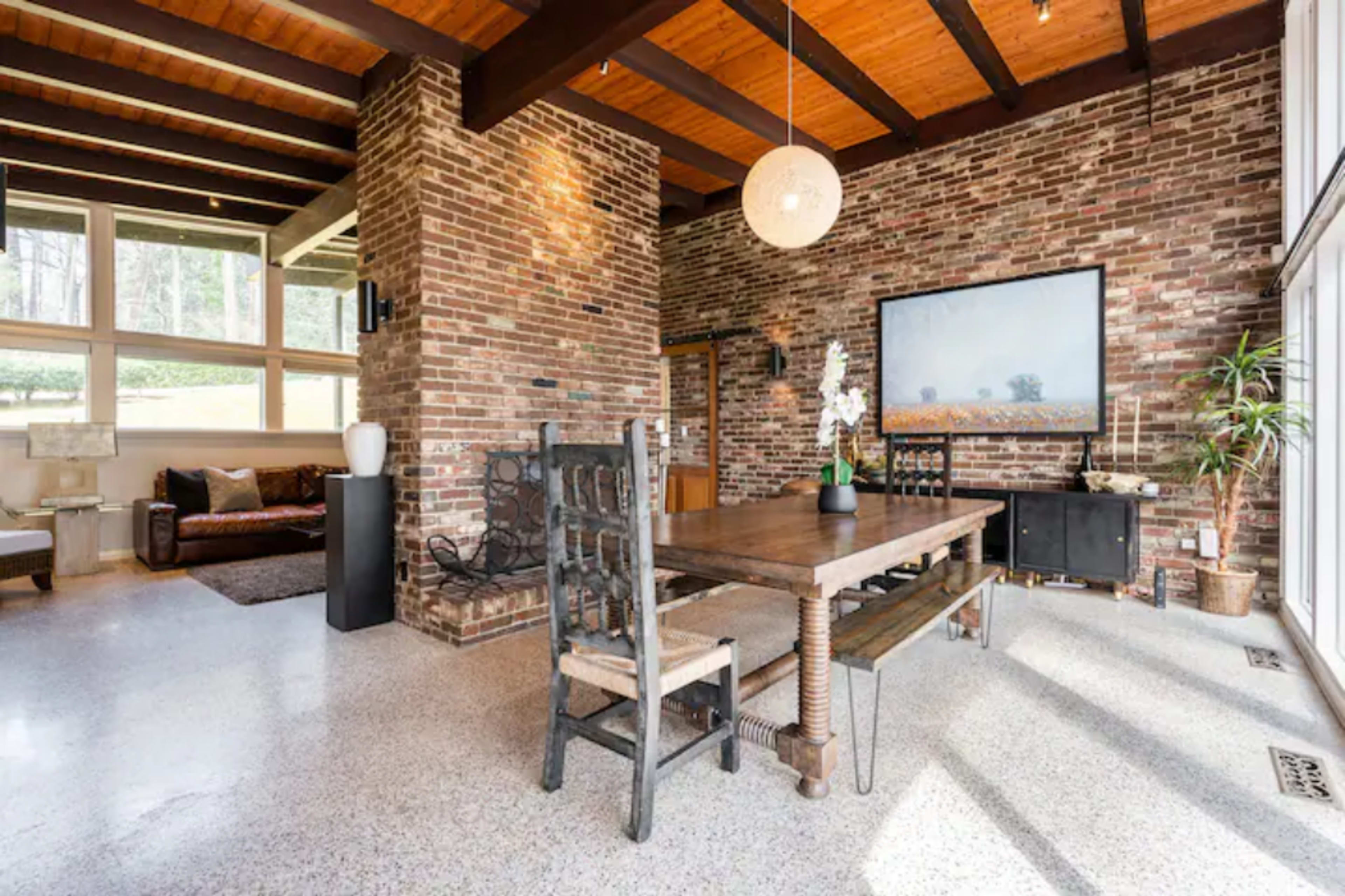 A modern living space featuring a dining table with rustic chairs, exposed brick walls, a large window, and wooden beams on the ceiling.