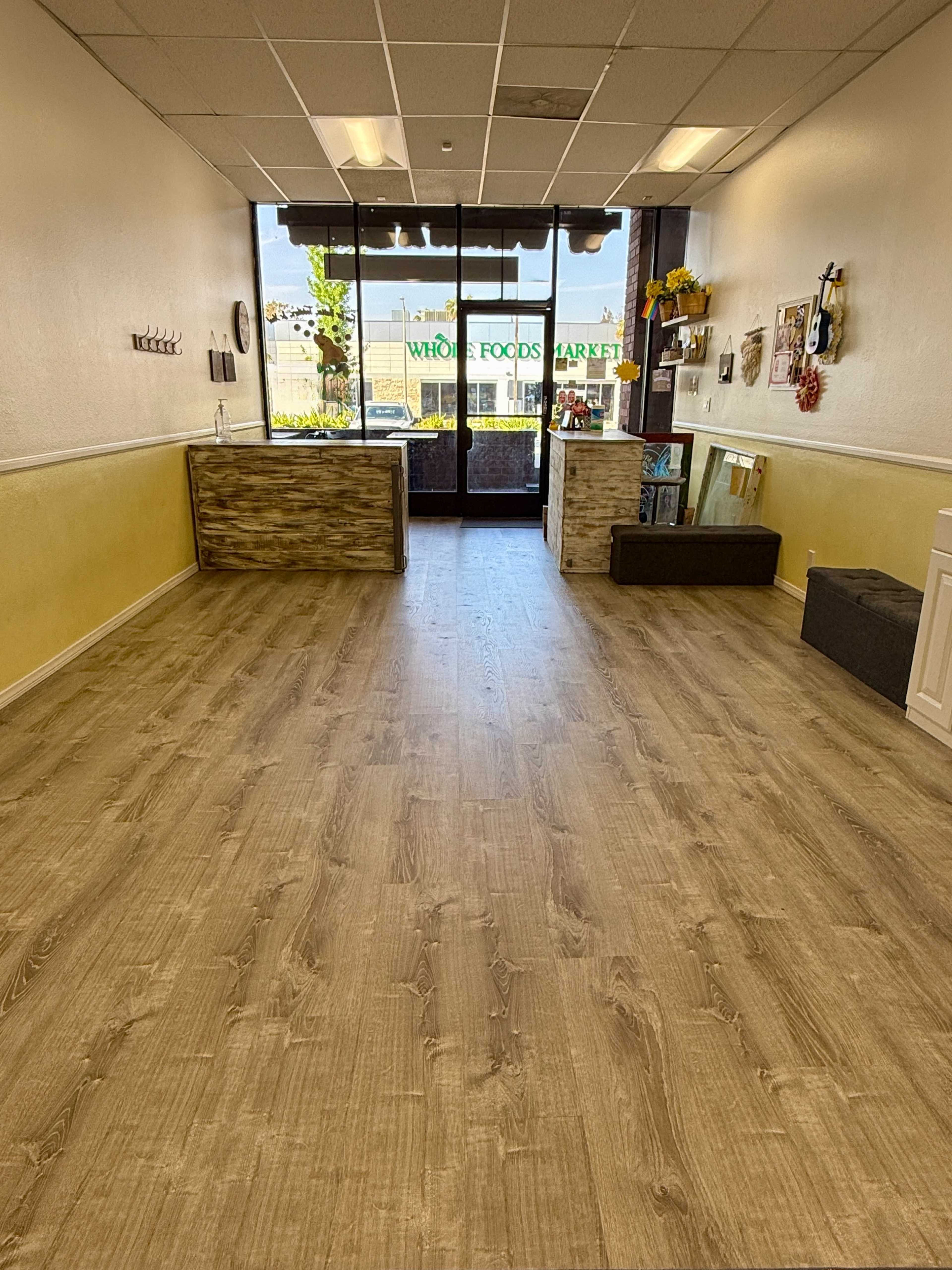 The image depicts a spacious, well-lit retail space with wooden flooring, a reception area on the left, and large windows facing a street with a view of a Whole Foods Market.