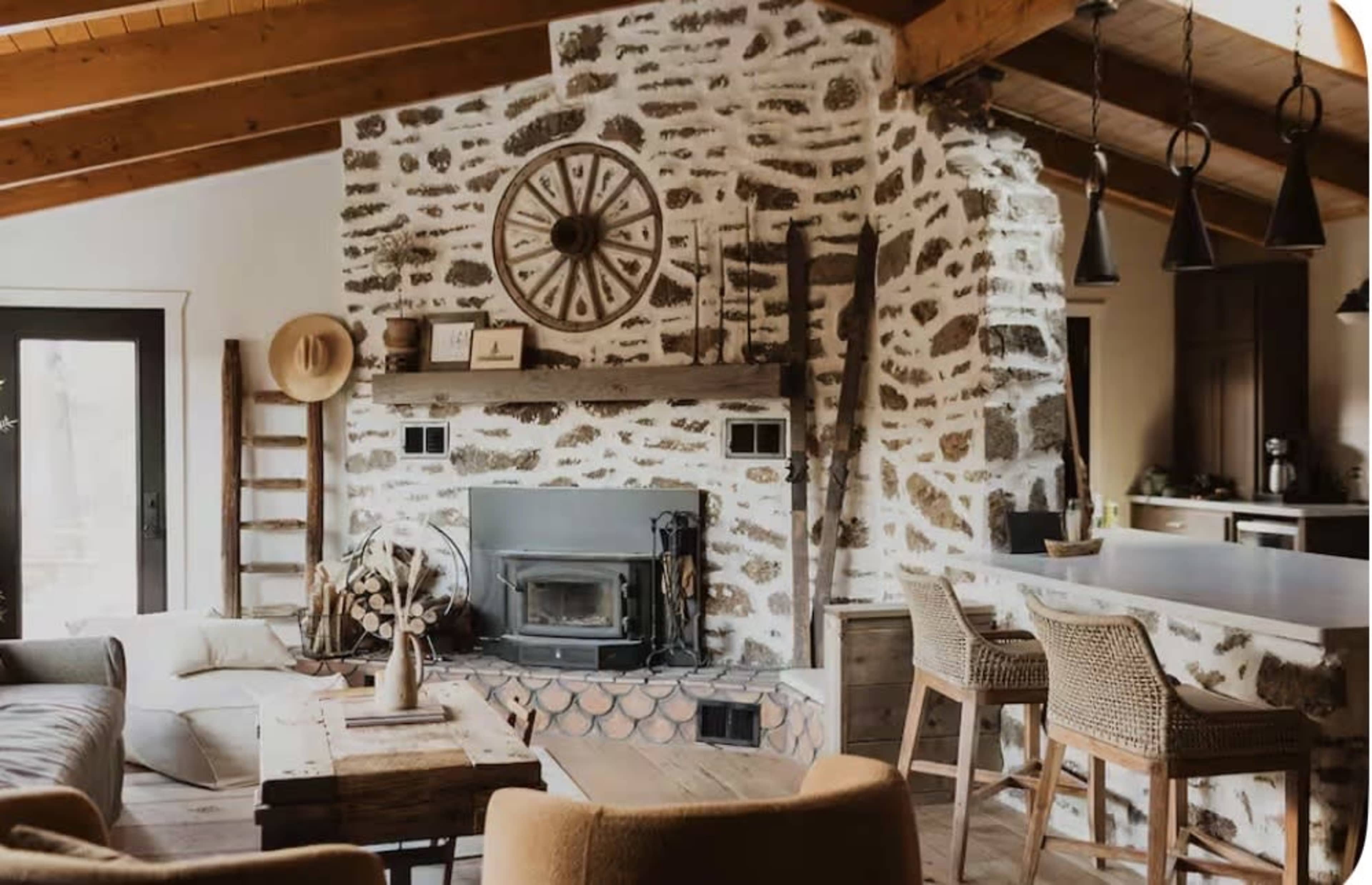 The image shows a cozy living room with a stone fireplace, wooden beams, and a mix of modern and rustic furniture.