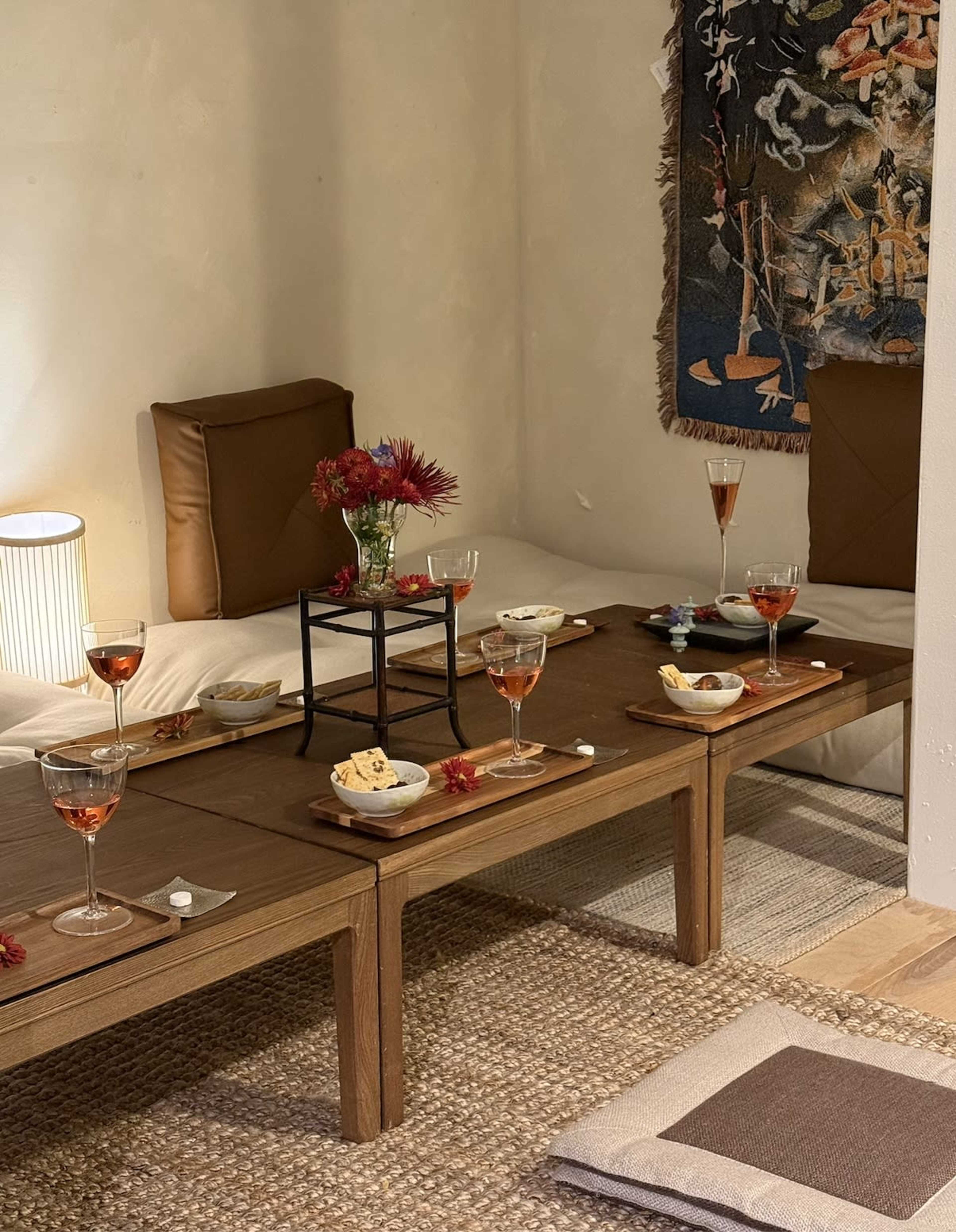 A low wooden table is set with glasses of pink drinks and small dishes, surrounded by floor seating in a warmly lit room adorned with a decorative wall hanging.