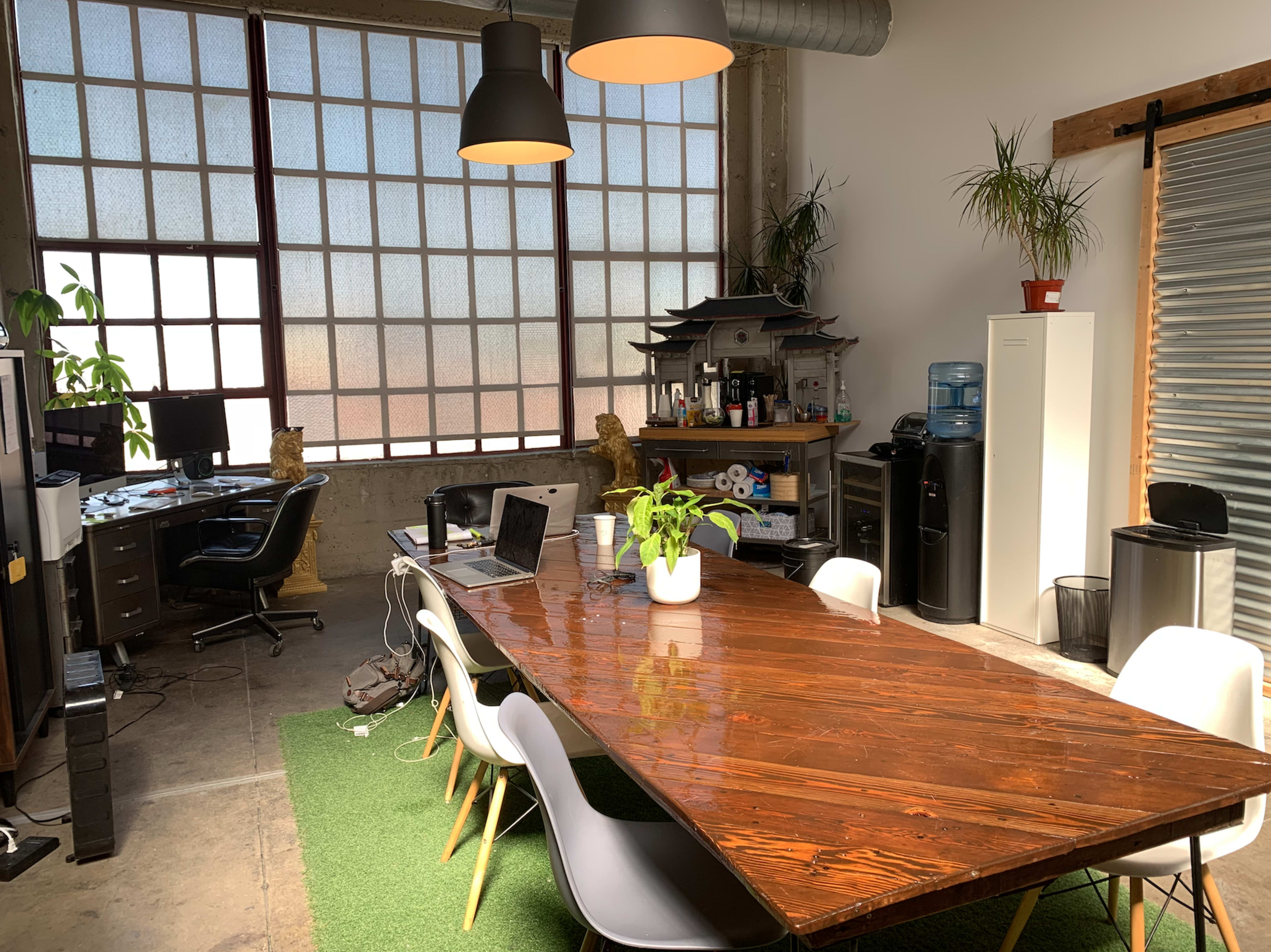 A large wooden conference table is surrounded by chairs in a bright office space featuring a desk with a computer, plants, and a water cooler.
