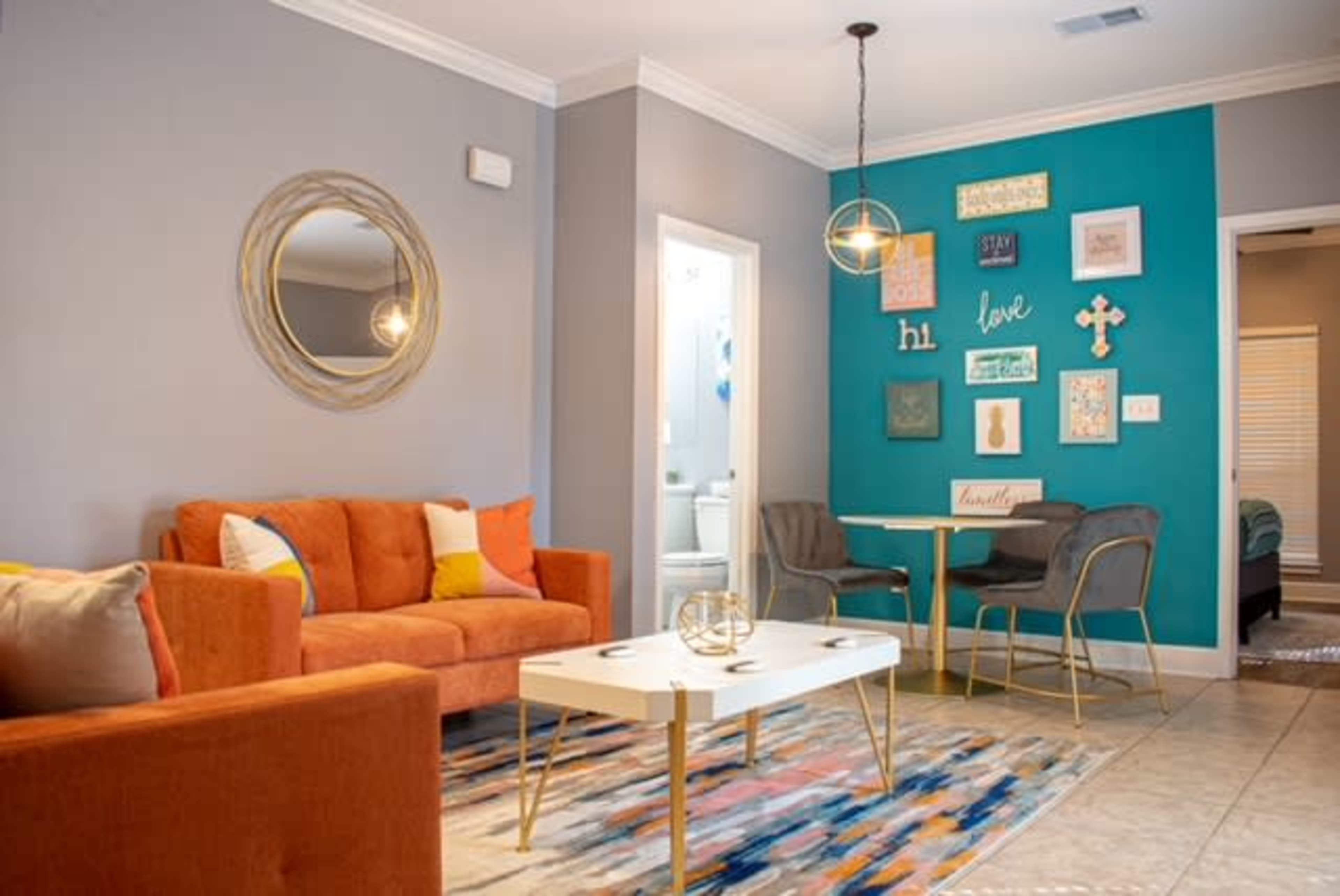 The image shows a living room with an orange couch, a round mirror, a white coffee table, and a dining area featuring teal walls adorned with framed decorations.