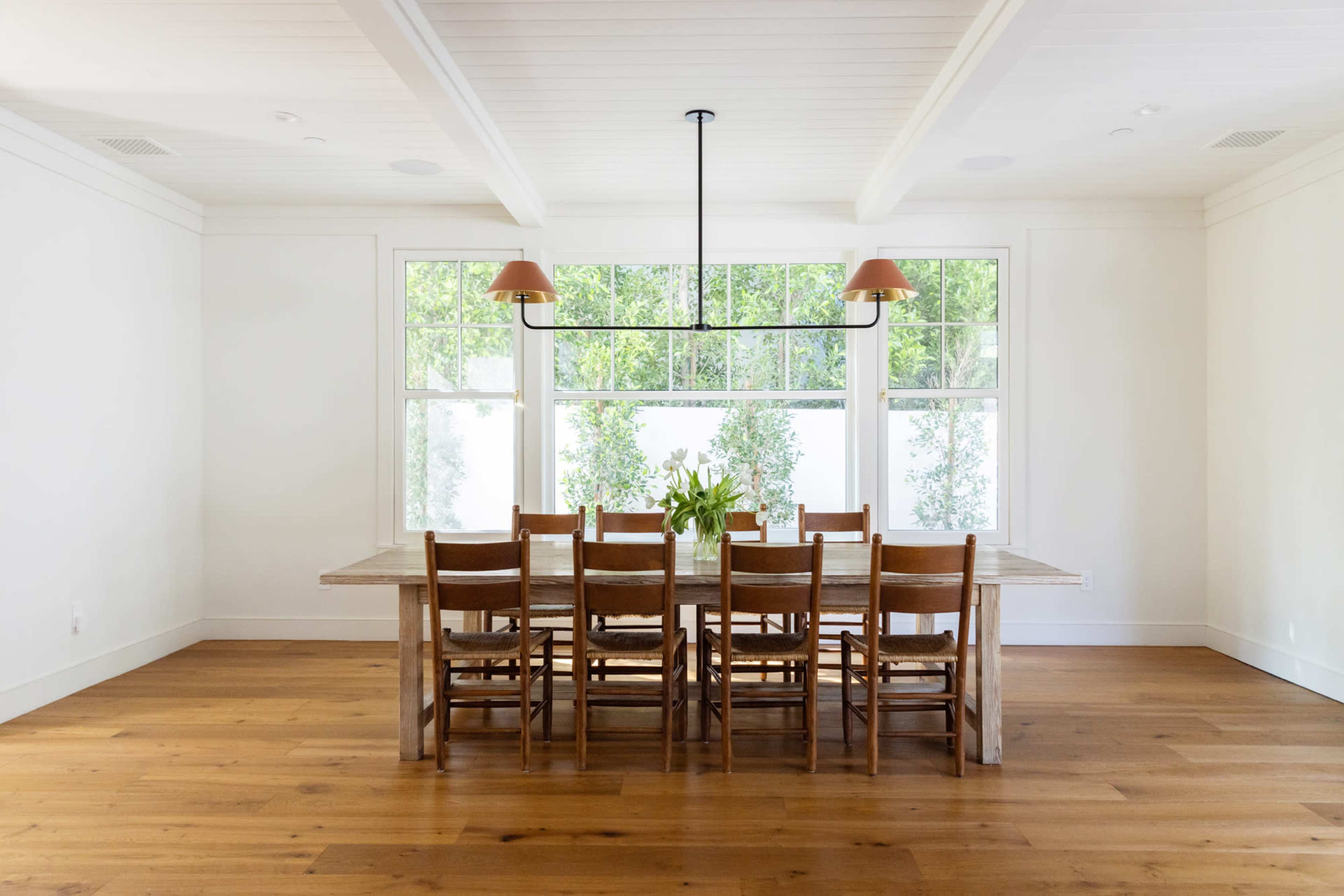 A long wooden dining table with eight chairs is positioned under a light fixture in a bright room featuring large windows.