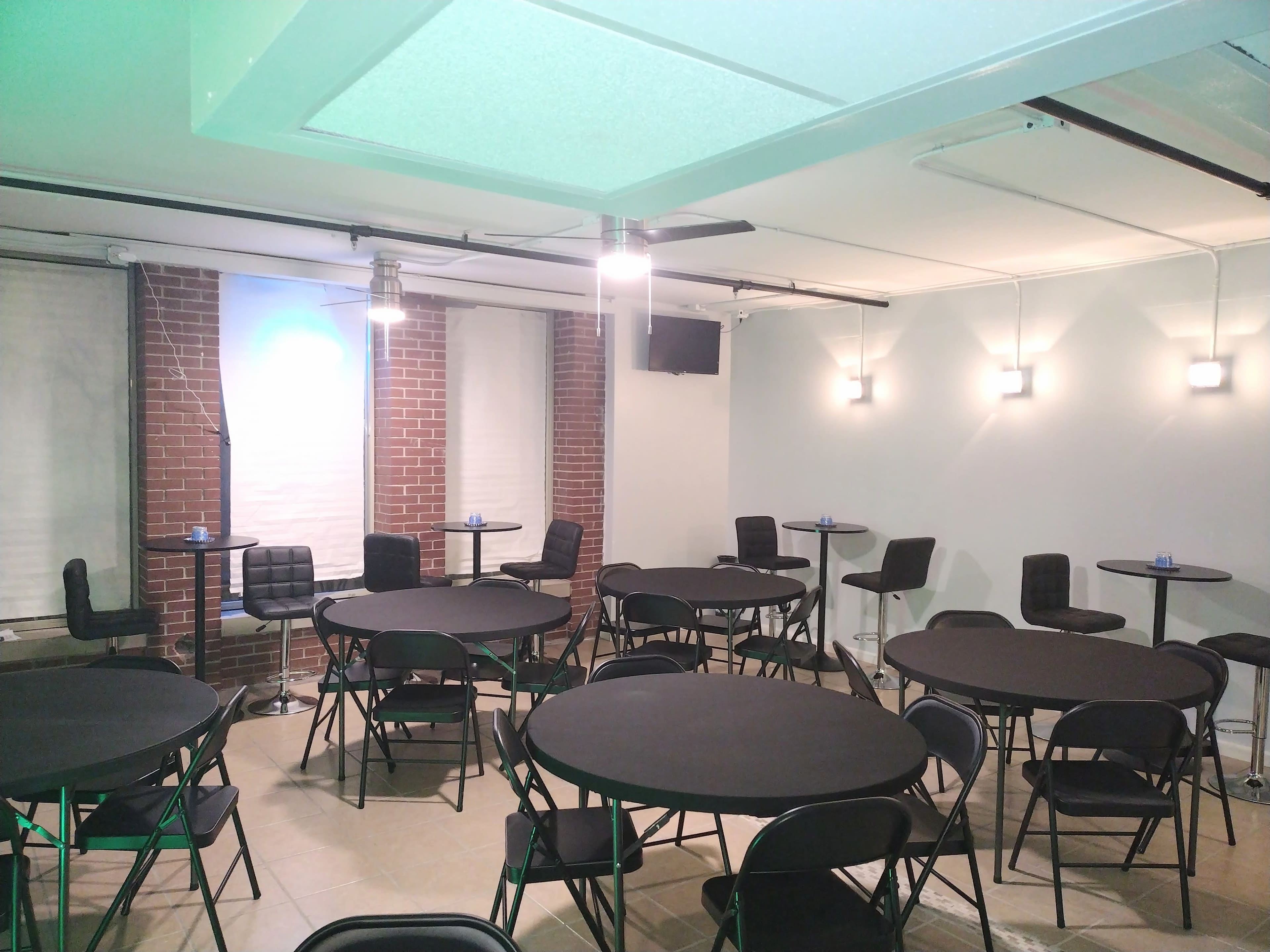 A room set up for an event, featuring several circular tables with black tablecloths surrounded by black chairs, and bright overhead lights illuminating the space.