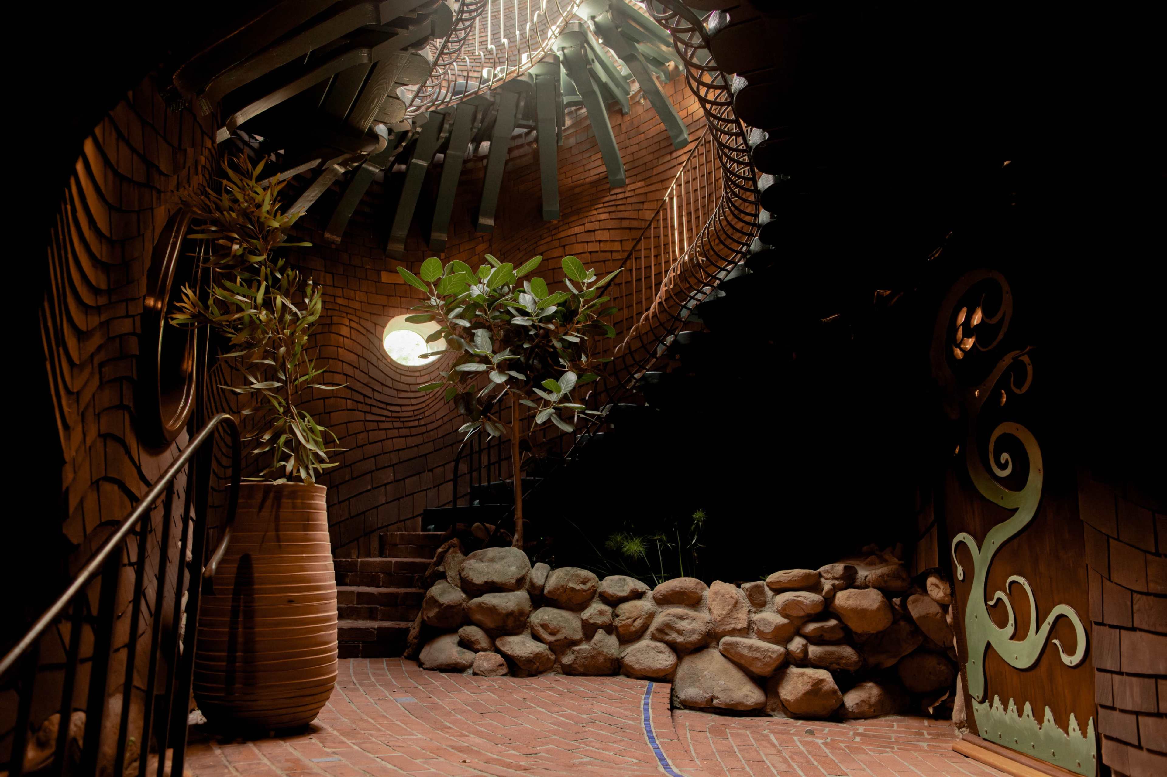 The image shows a surreal interior space featuring a winding staircase surrounded by a unique, textured wall, with plants and rocks integrated into the design.