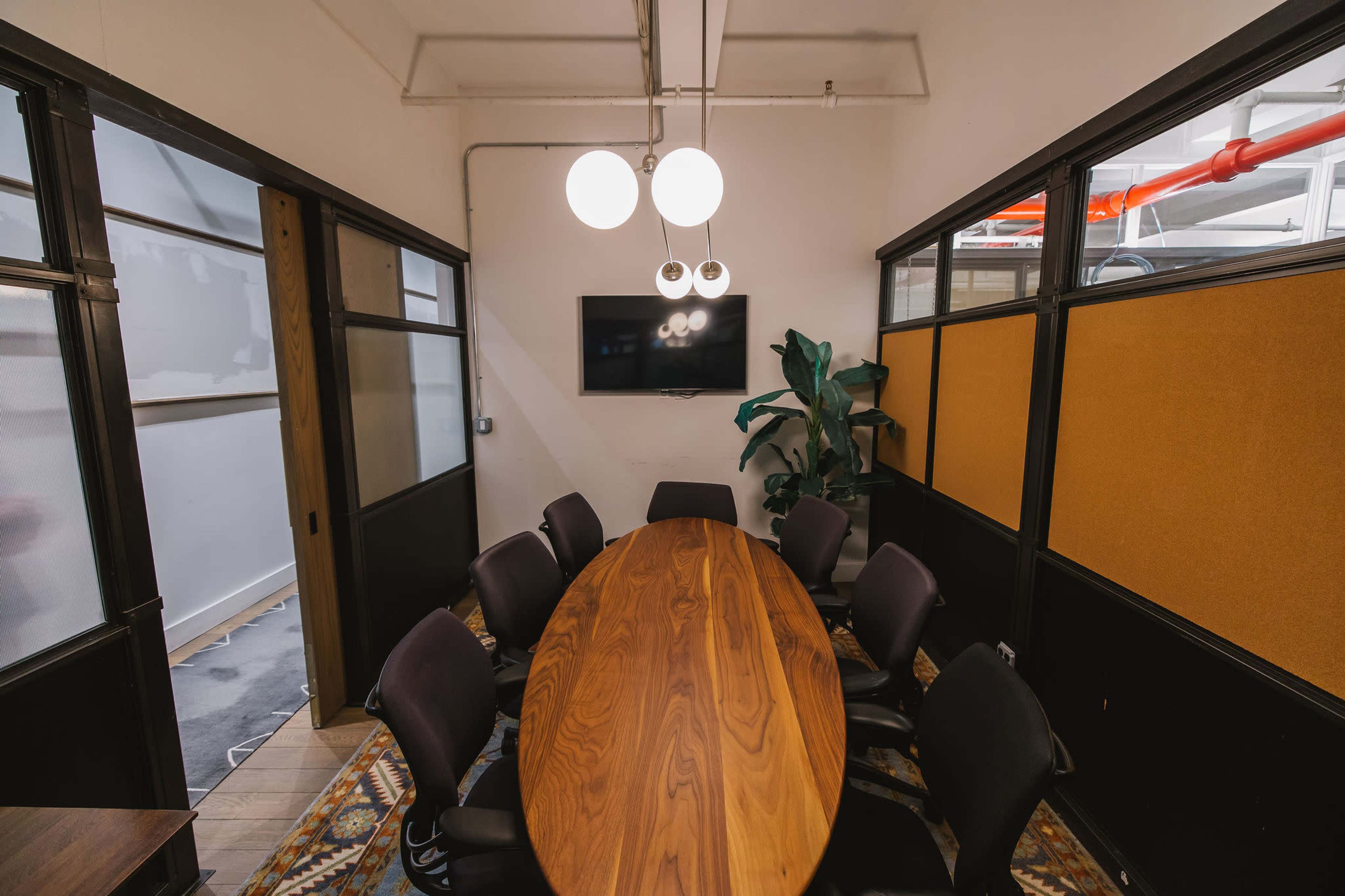 A modern conference room features an oval wooden table surrounded by black chairs, with a wall-mounted screen and pendant lighting overhead.