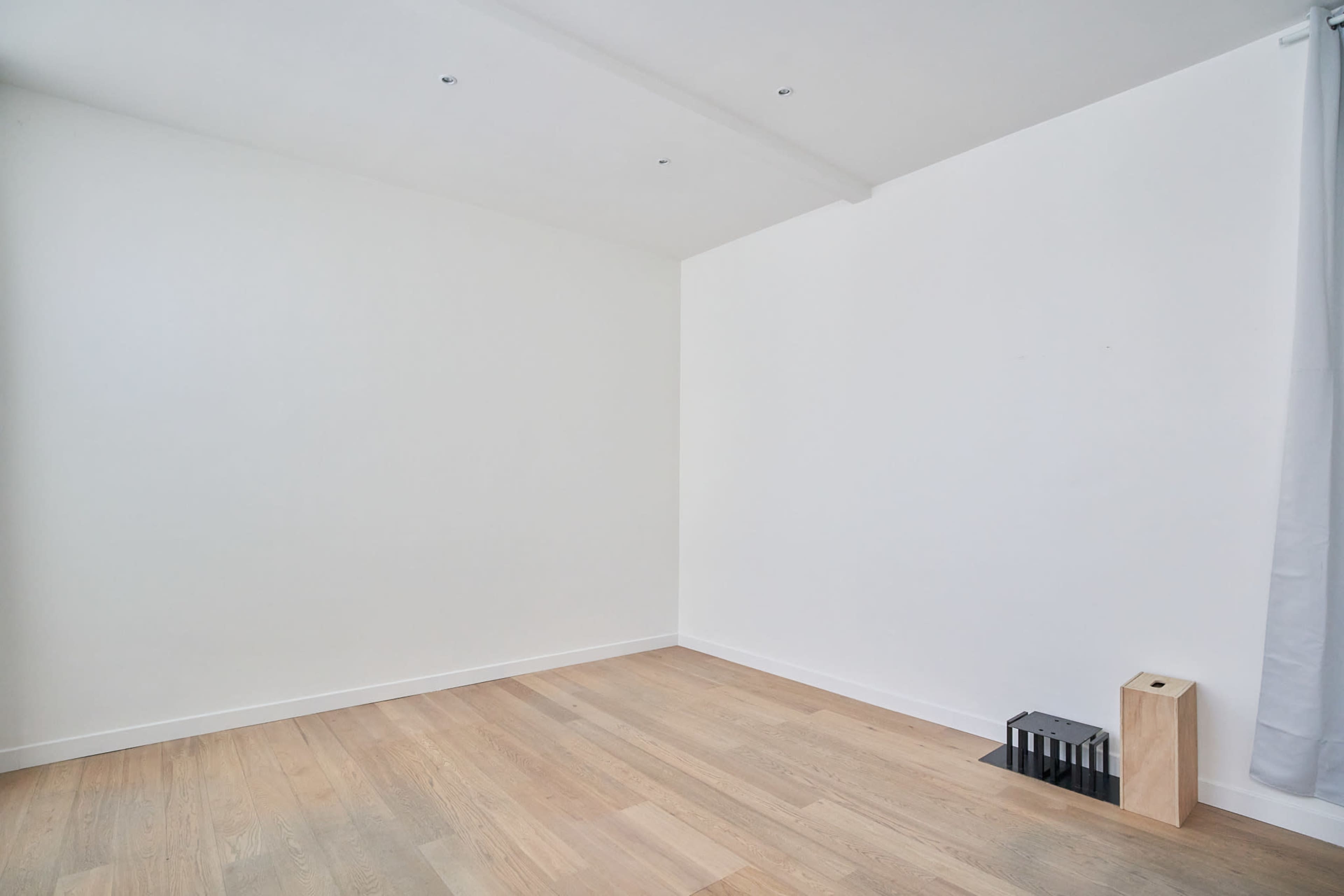 An empty room with white walls, wooden flooring, and minimal furnishings including a small wooden block and a black storage unit against one wall.